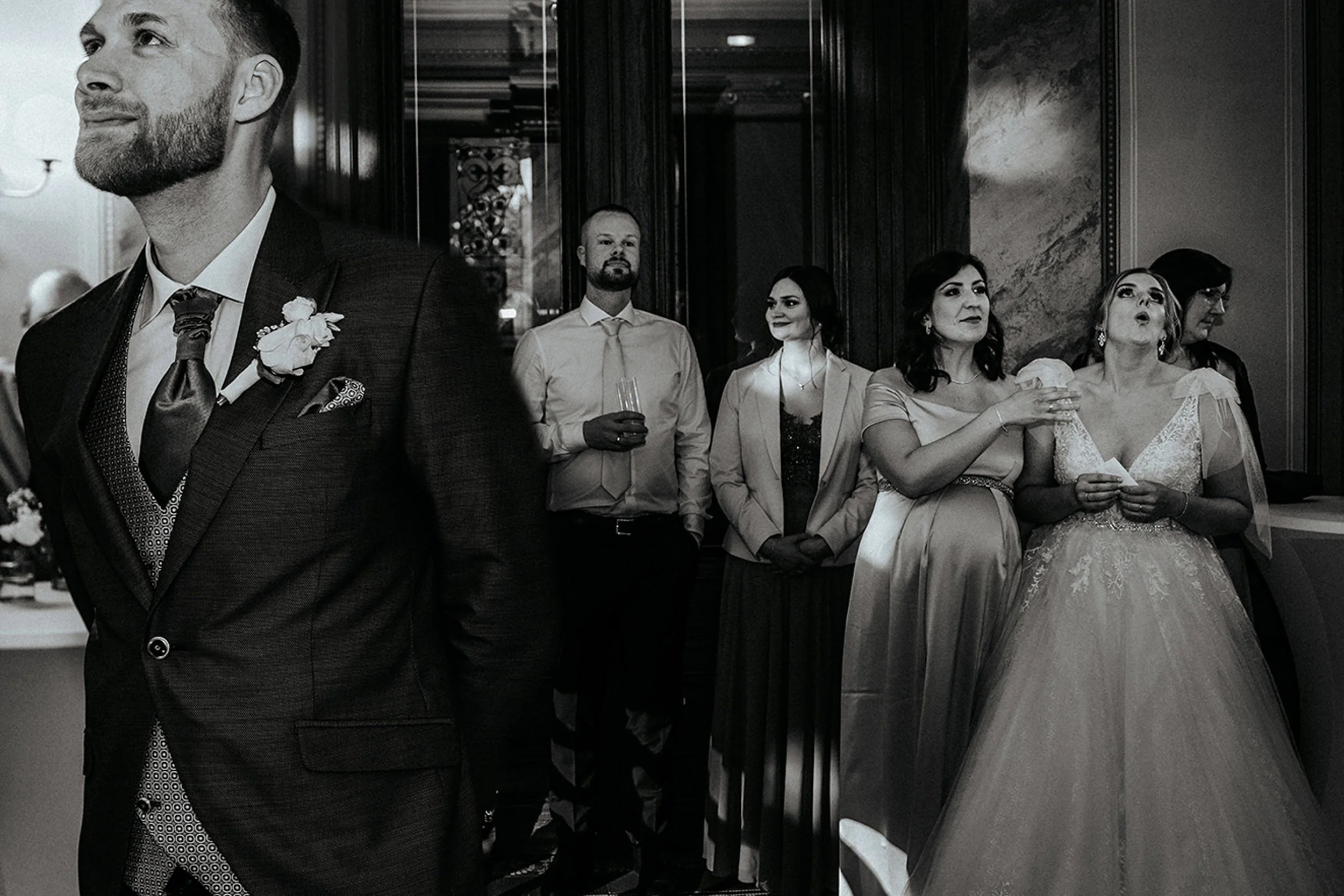 Eine Gruppe von Menschen bei einer formellen Veranstaltung. Im Vordergrund ein Mann in Anzug und Fliege, im Hintergrund mehrere Männer und Frauen, die auf einer Party oder Hochzeit zu sehen sind.