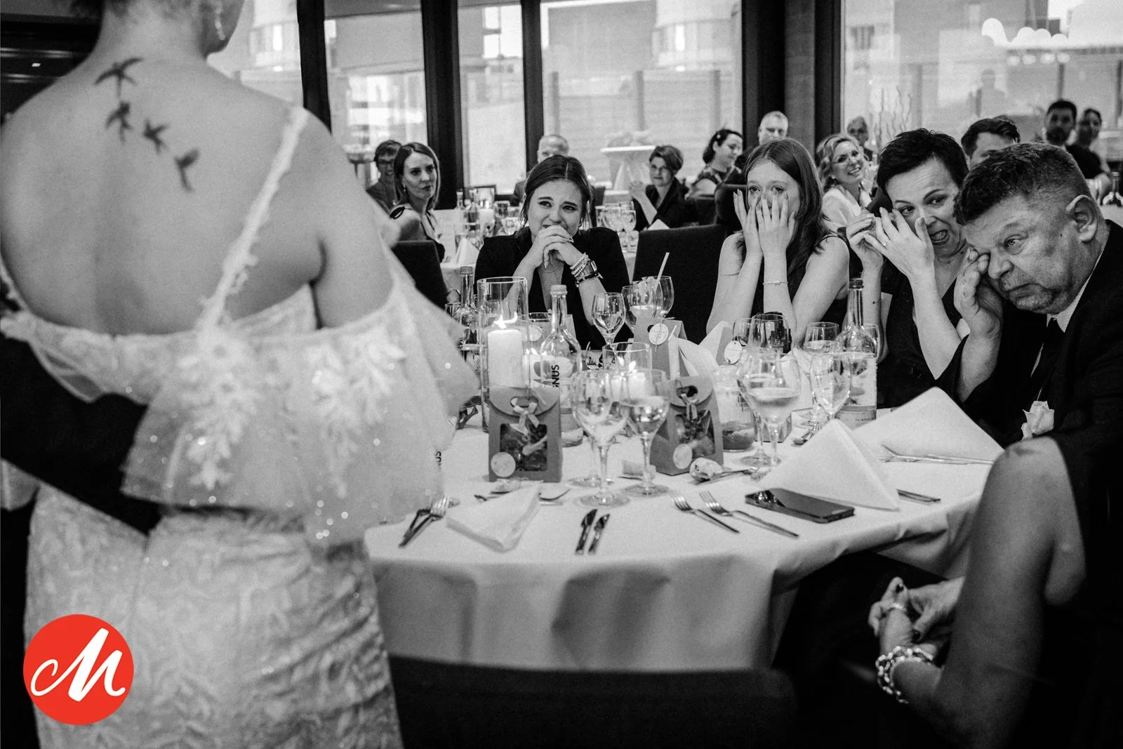 Frau in Brautkleid bei einer Hochzeit spricht vor einer Tafel voller Gäste, die emotional reagieren.