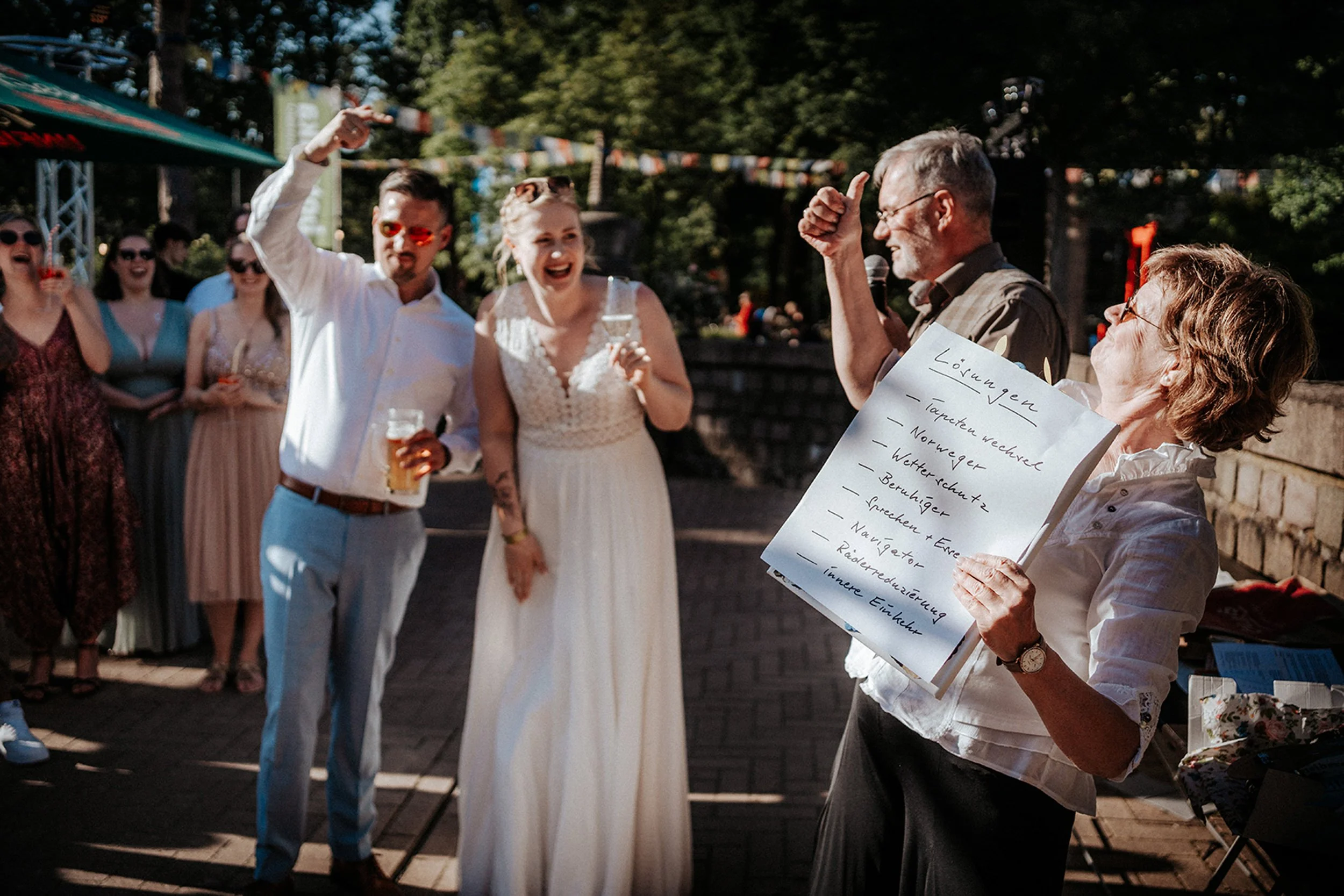 Eine Gruppe von Menschen, darunter ein Brautpaar, steht im Freien, während eine Frau ein Whiteboard mit einer Liste hält. Es sieht nach einer Hochzeitsfeier aus, geladene Gäste feiern und lachen in der Sonne.