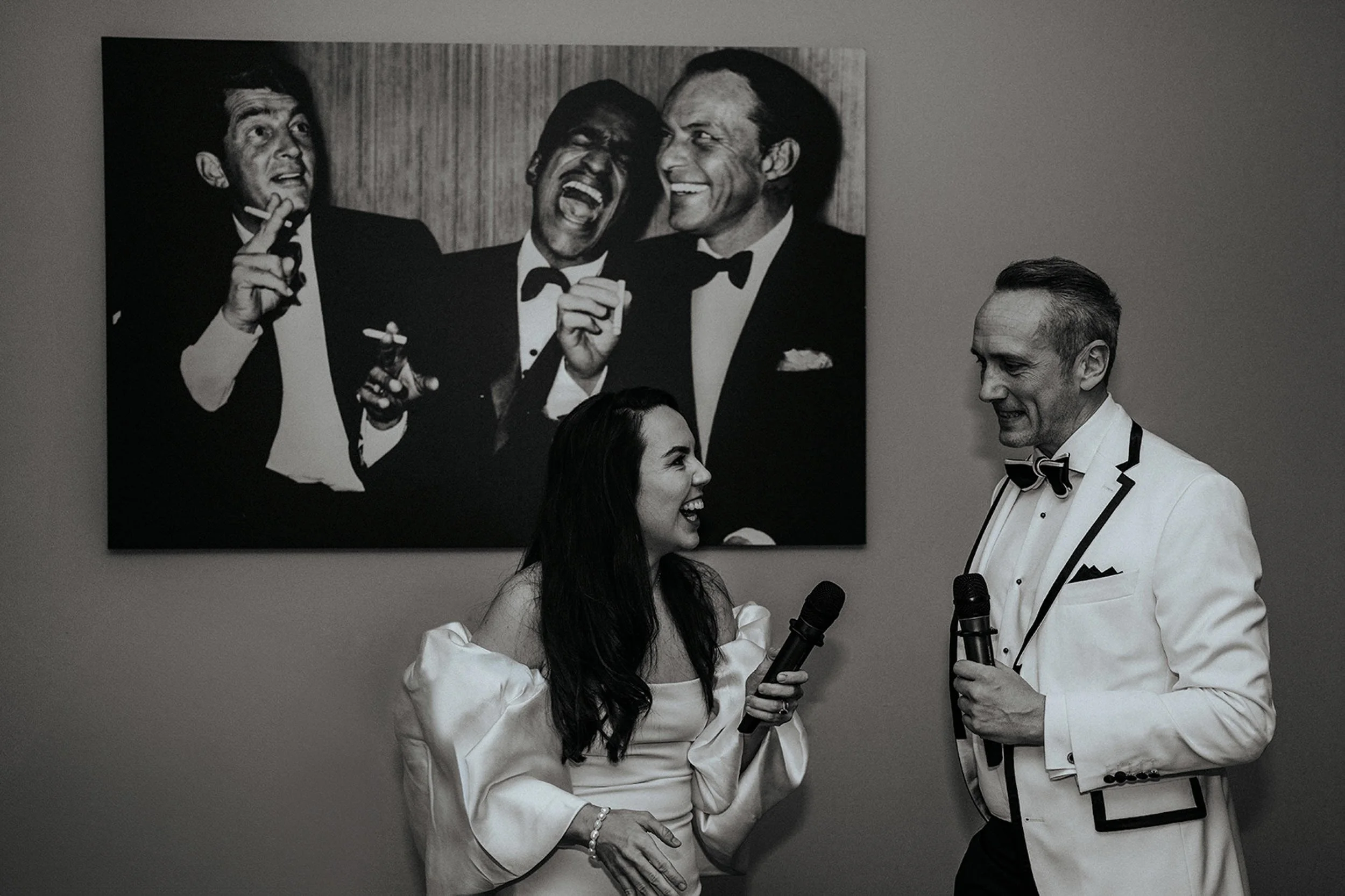 Schwarz-Weiß-Fotografie von drei Menschen in formeller Kleidung, die sich auf einer Party oder Veranstaltung amüsieren, mit einem Poster im Hintergrund, das drei lachende Männer in Anzügen zeigt.