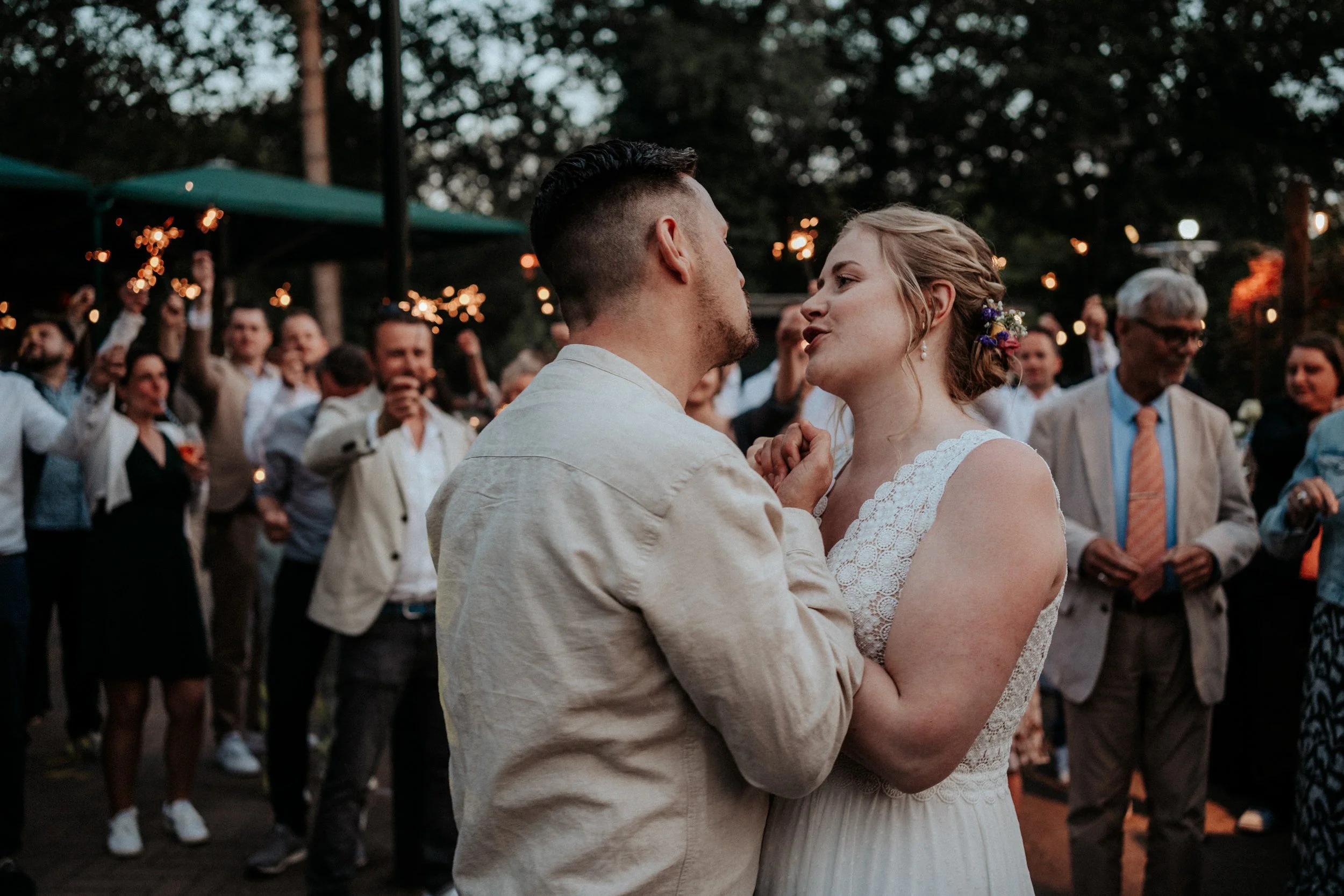 Ein Paar tanzt bei einer Hochzeit im Freien, umgeben von fröhlichen Gästen, mit Lichterketten im Hintergrund.