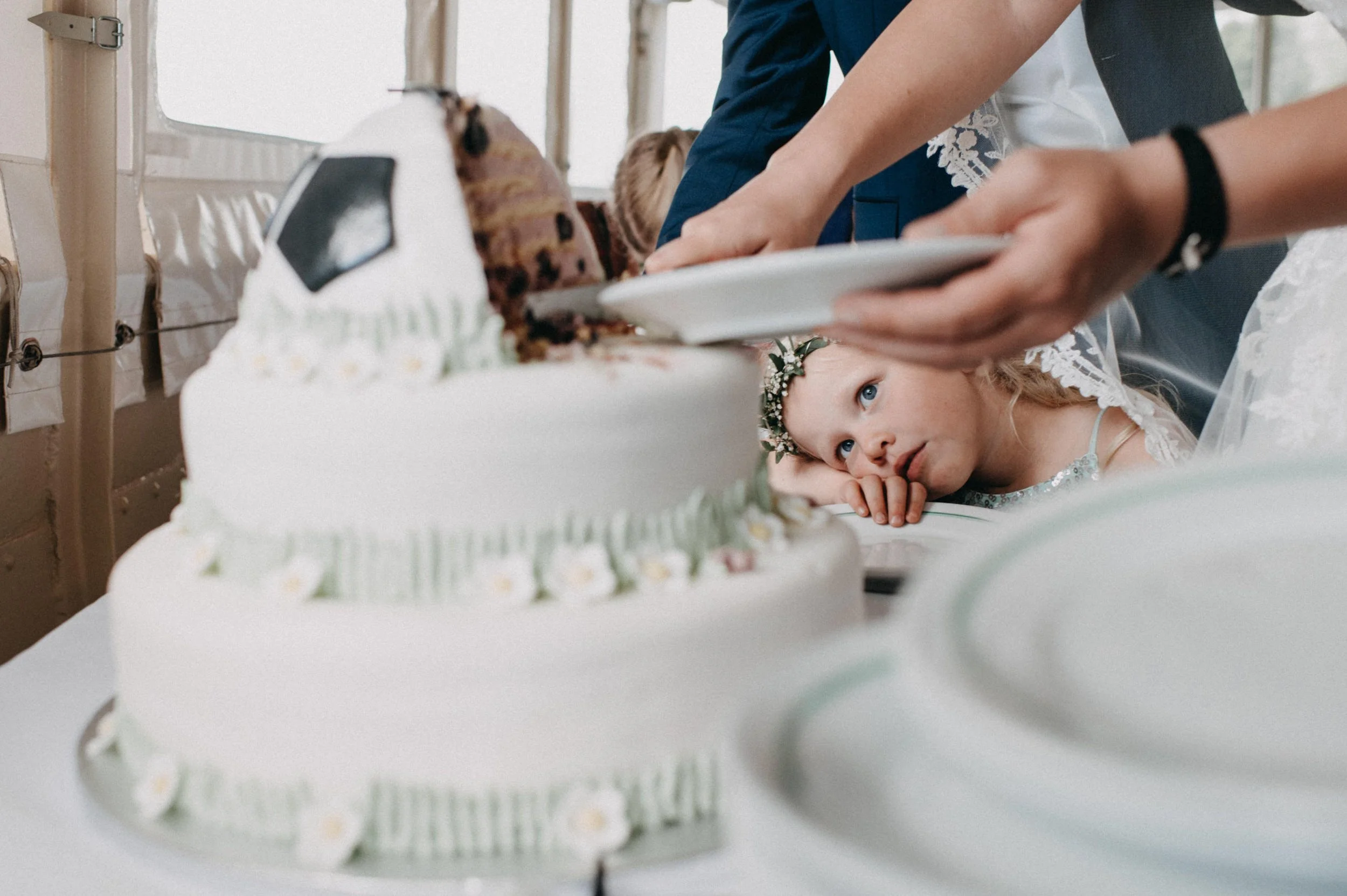 Ein kleines Mädchen mit Blumenkranz im Haar liegt auf dem Tisch und schaut auf eine Hochzeitstorte, während Menschen die Torte schneiden.
