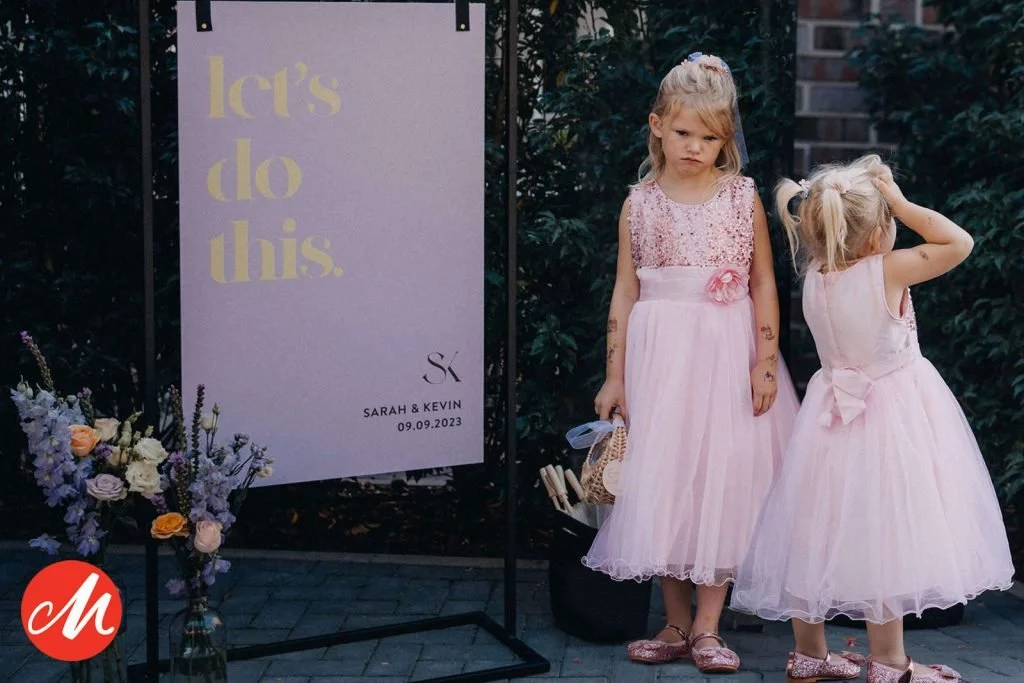 Zwei kleine Mädchen in pinken Tüllkleidern stehen neben einem Begrüßungsschild mit der Aufschrift "Let's do this" bei einer Hochzeit am 09.09.2023 für Sarah & Kevin.