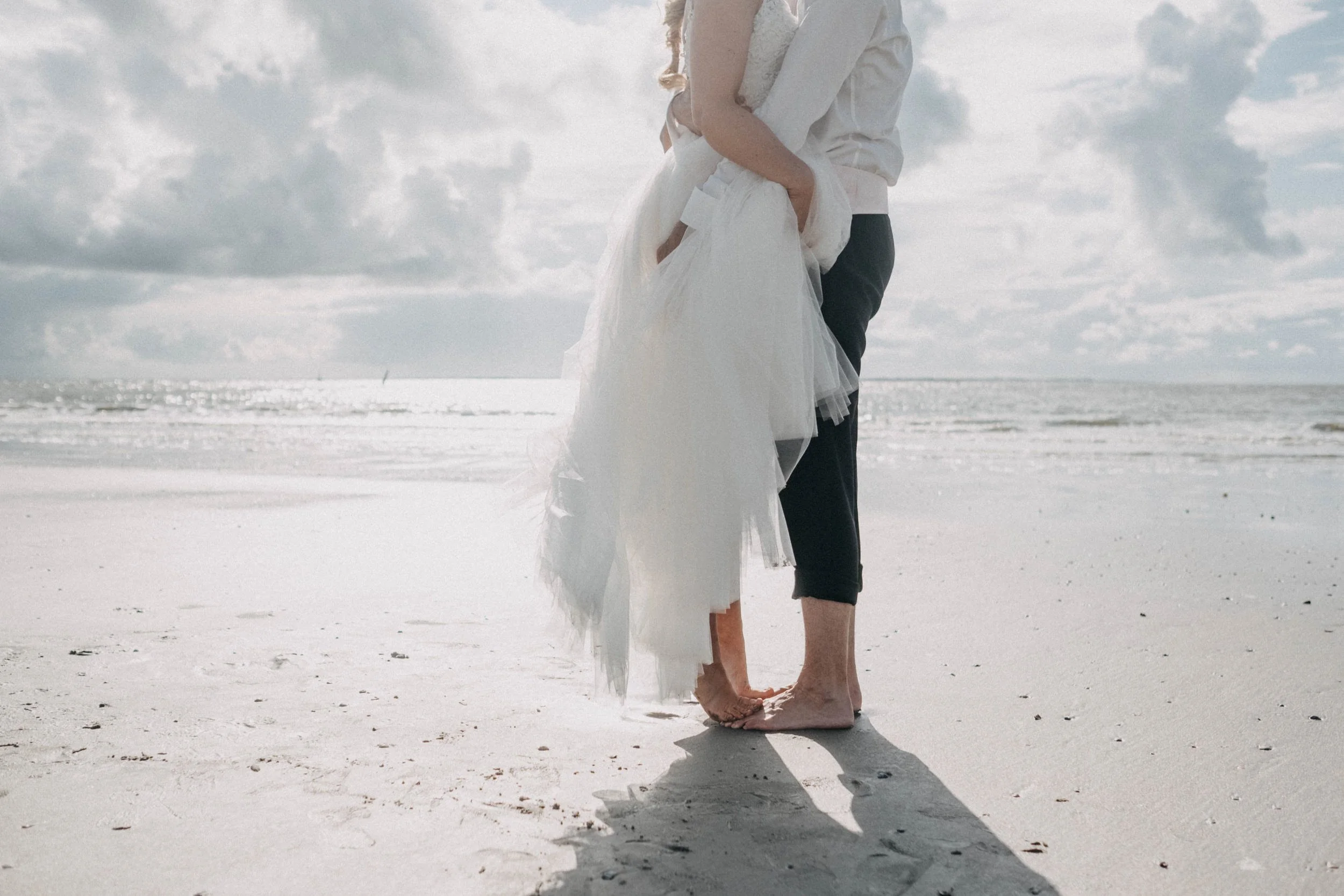 Ein Paar steht barfuß im Sand am Strand, elterliche Umarmung, Himmel mit Wolken, Meer im Hintergrund.