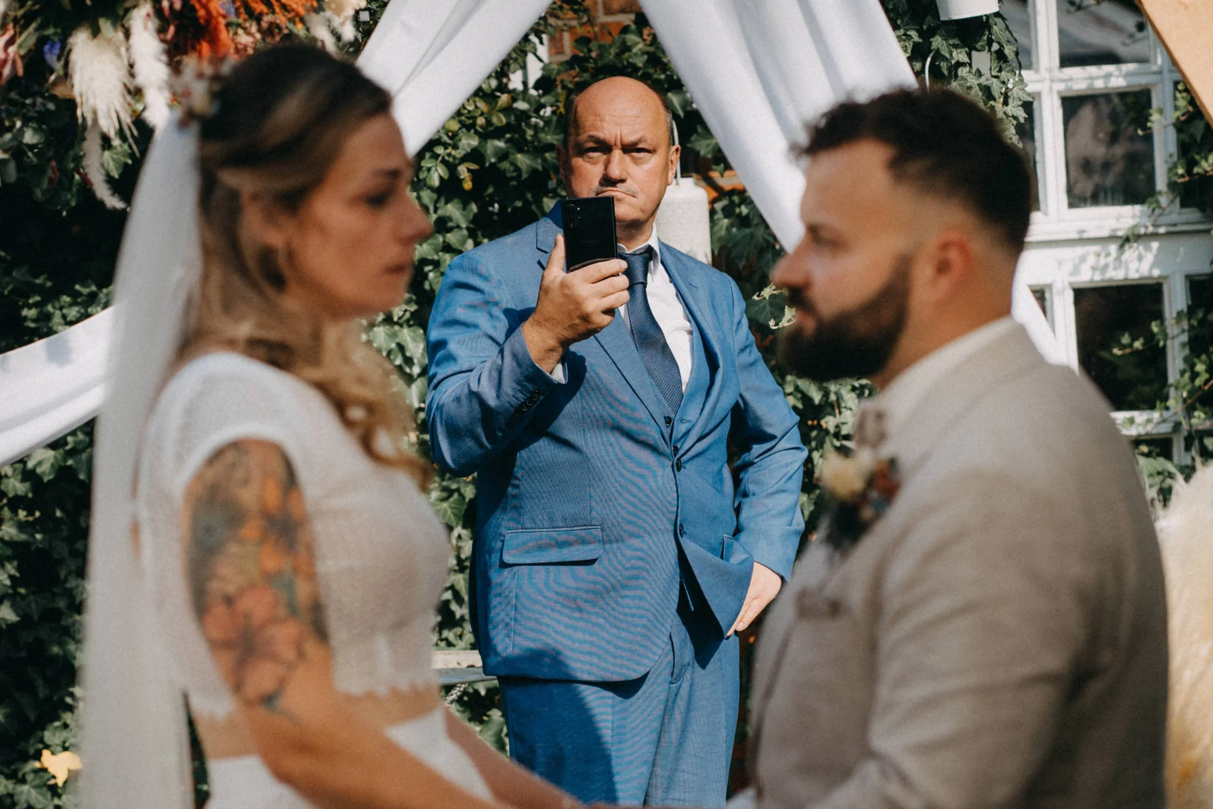 Hochzeitsszene mit einem Mann in blauer Anzug hinter einem Brautpaar, das sich gegen einen weißen Bogen mit grünen Pflanzen im Hintergrund freut. Der Mann schaut ernst, während die Braut und der Bräutigam lachend in der Nähe sind.