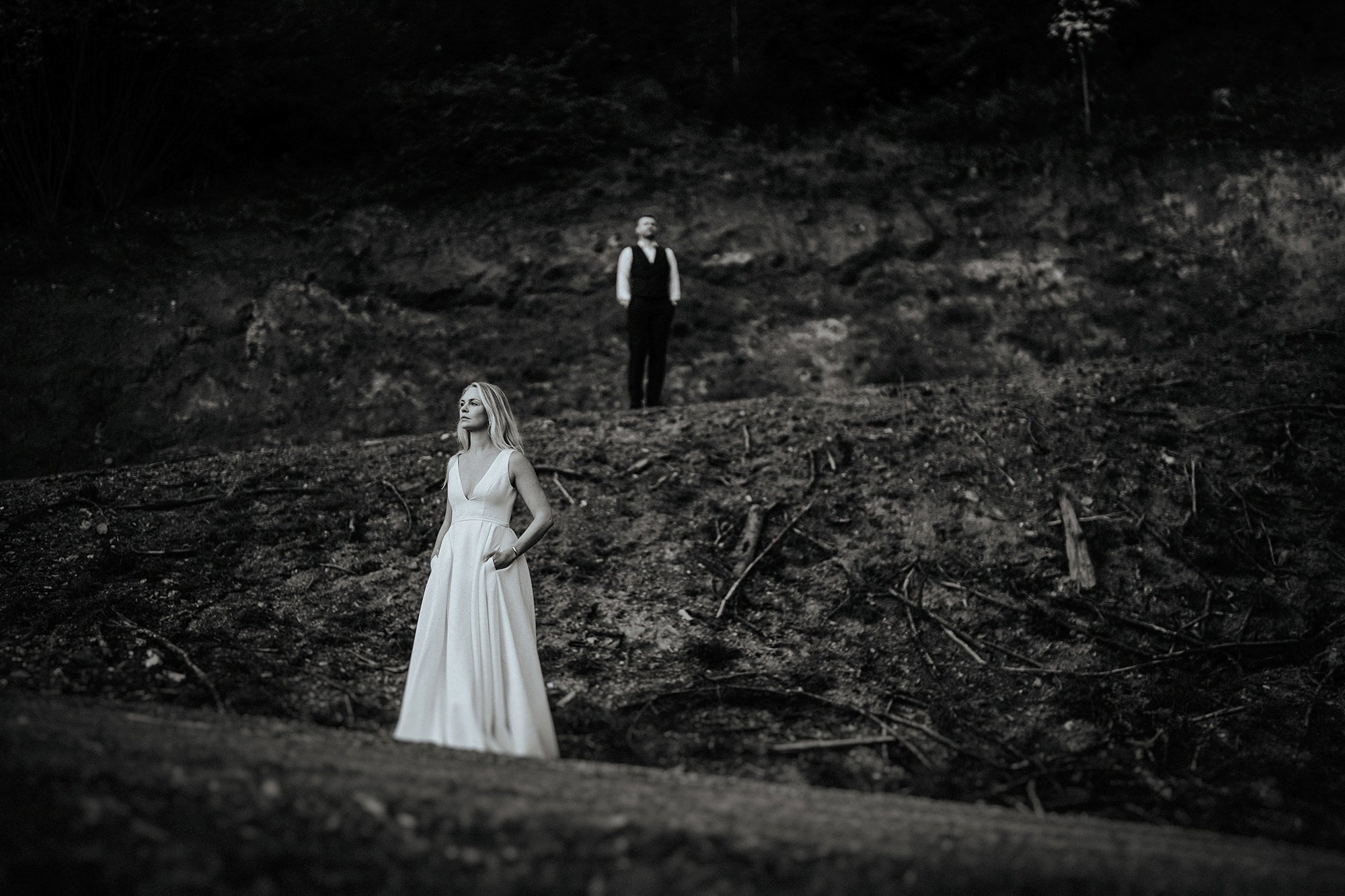 Ein Schwarz-Weiß-Foto von zwei Menschen in formeller Kleidung in einer unberührten, dunklen, unebenen Landschaft, die an einen Wald oder ein verlassenes Gelände erinnert. Eine Frau in einem weißen Kleid steht im Vordergrund, während eine Person im sc