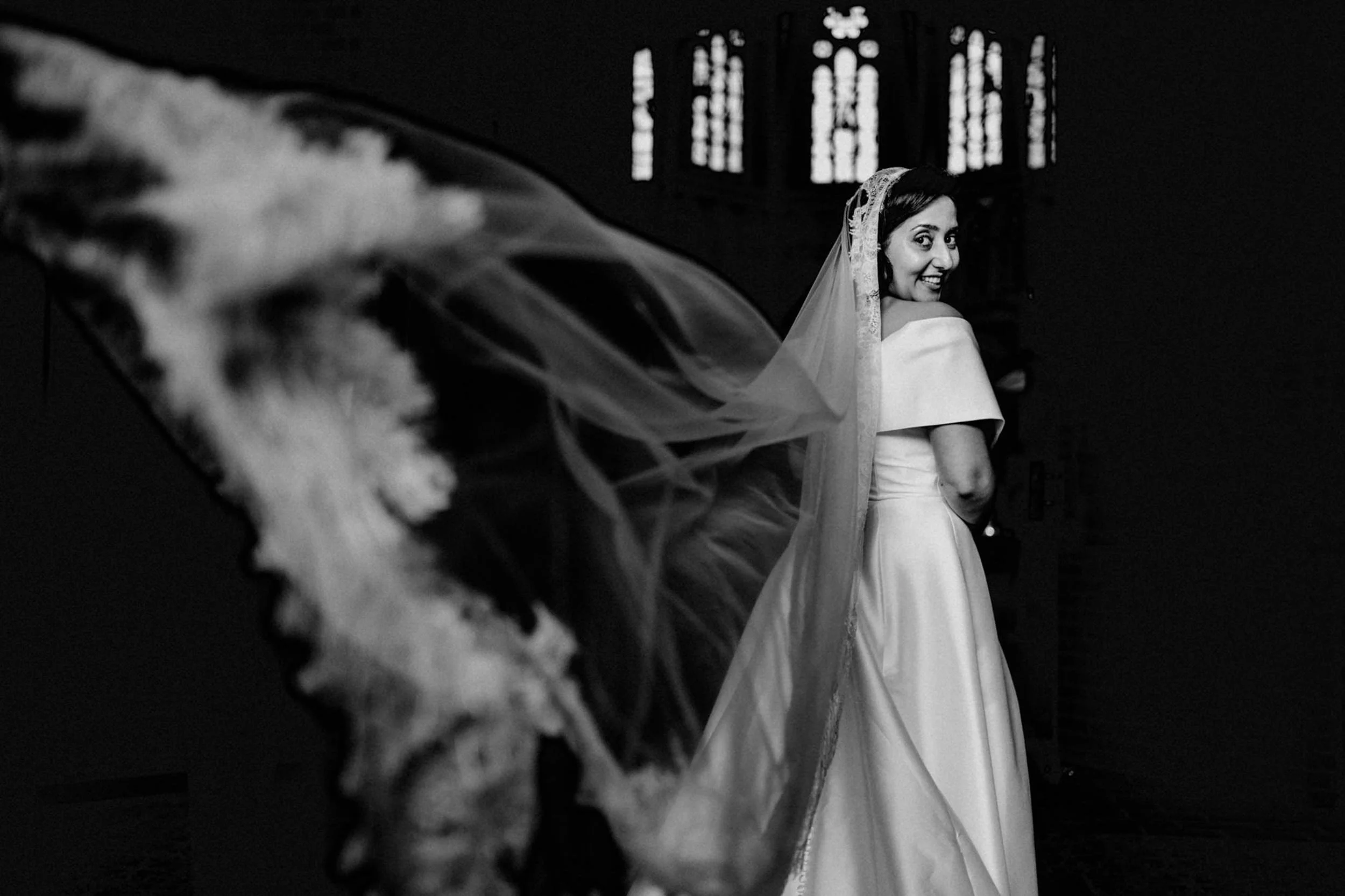 Schwarz-weiß-Foto einer lächelnden Braut in einem Brautkleid und Schleier, im Hintergrund ein Kirchenfenster.