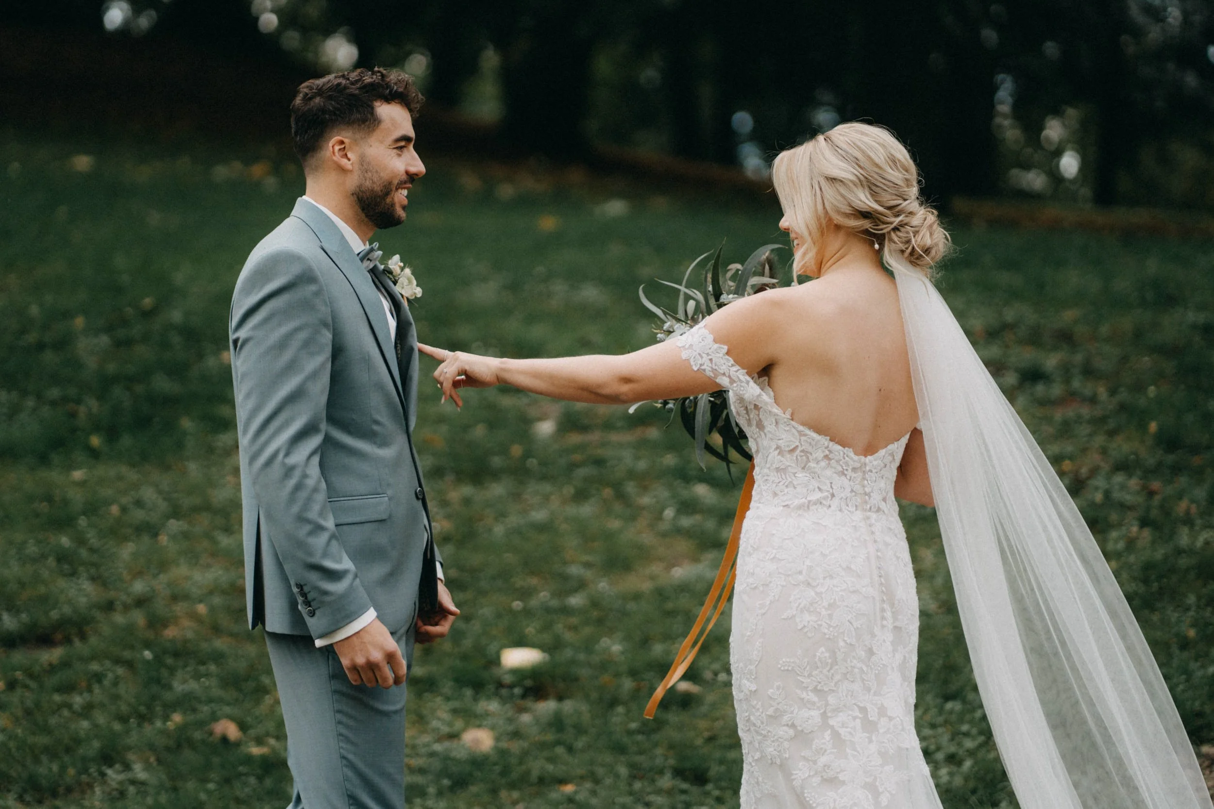 Ein Brautpaar während einer Hochzeit im Freien, der Bräutigam trägt einen grauen Anzug, die Braut trägt ein weißes, elegante Hochzeitskleid mit Spitzen. Sie erscheinen glücklich und verliebt.