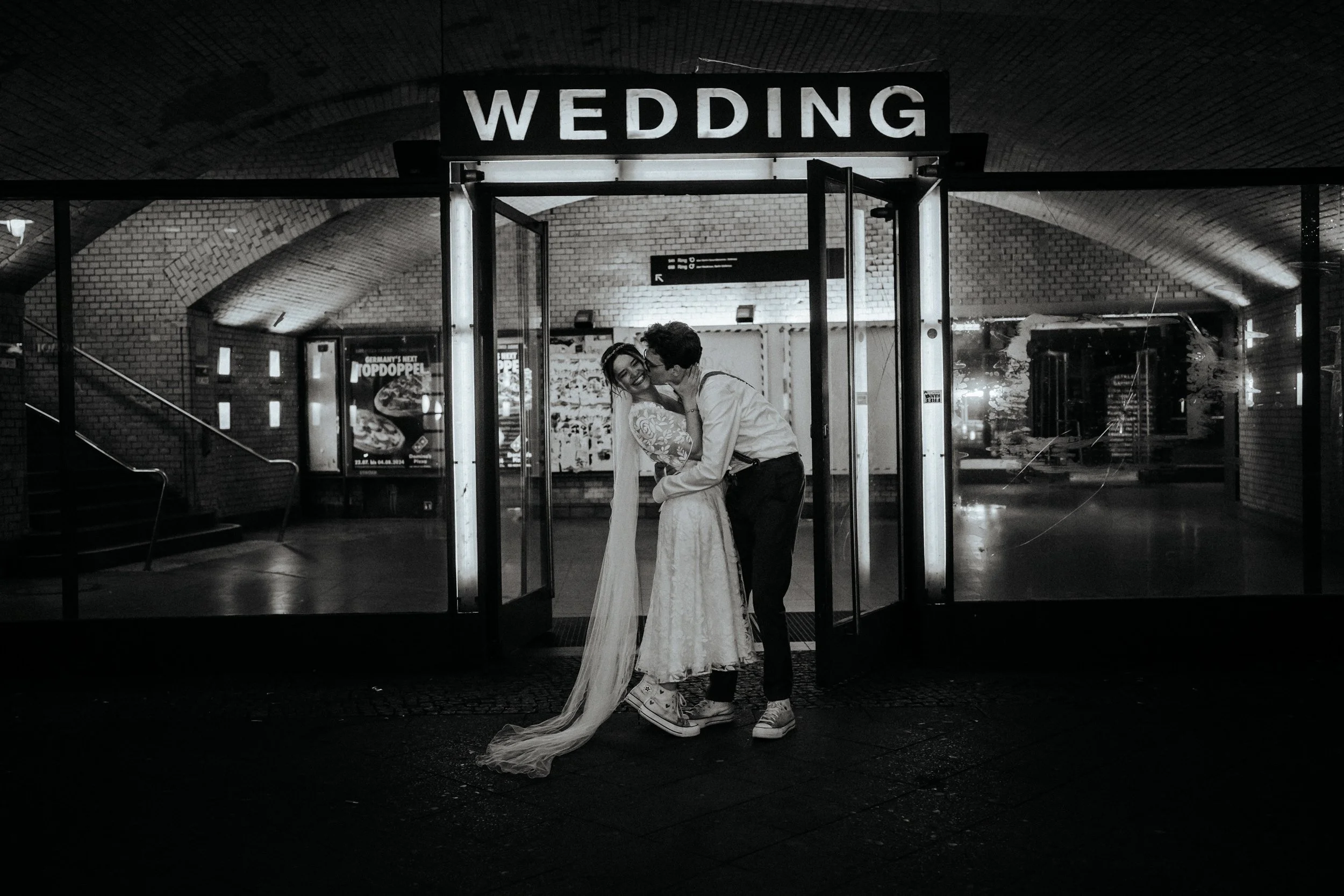 Ein Brautpaar im Schlosshof bei Nacht, das sich küsst, während sie vor einem Eingang mit einem Schild "Wedding" steht.
