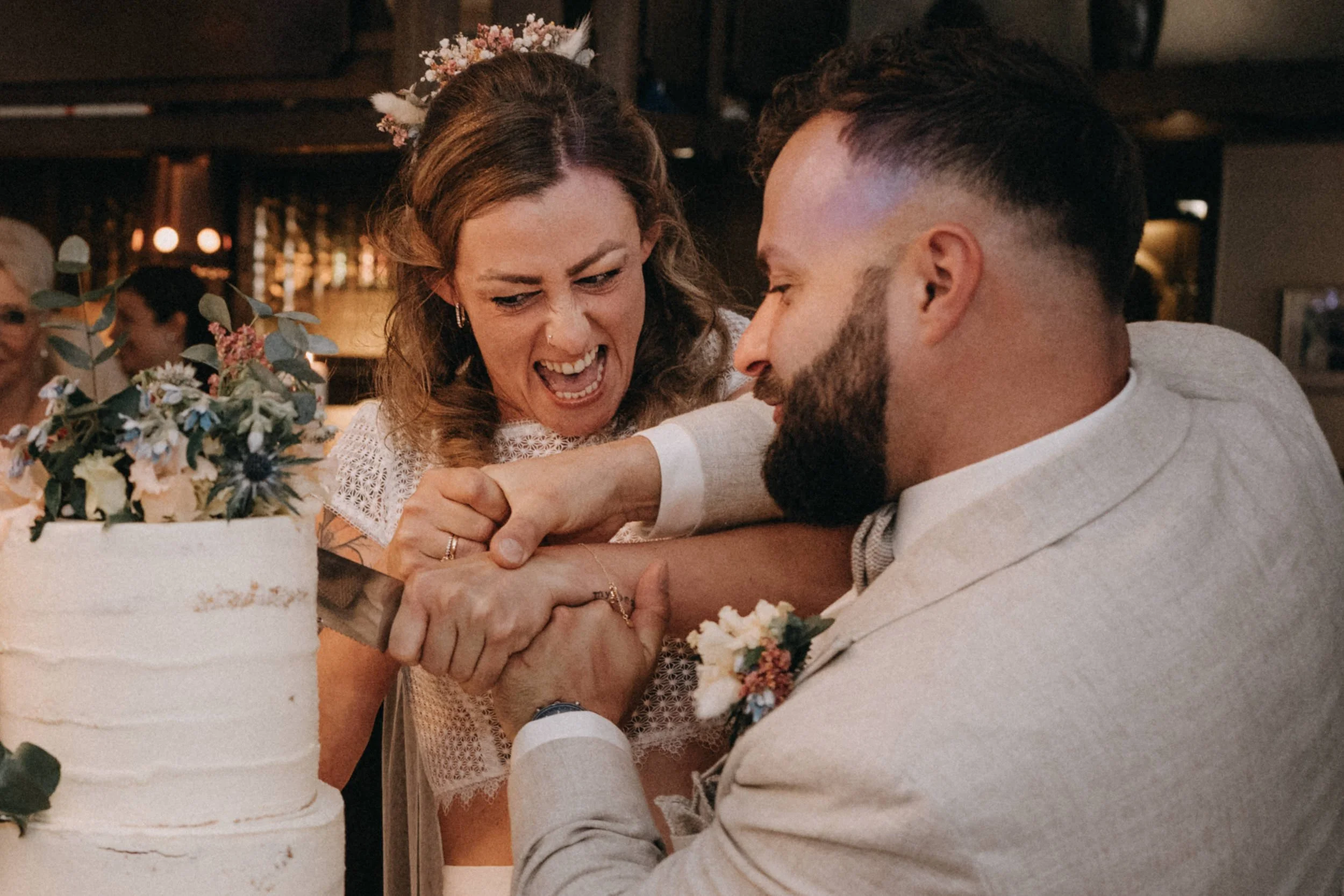 Ein glückliches Paar bei einer Hochzeit, umarmt sich mit verschwindender Gesellschaft im Hintergrund, auf einem festlichen Tisch mit Blumen und einem weißen Hochzeitstorte.