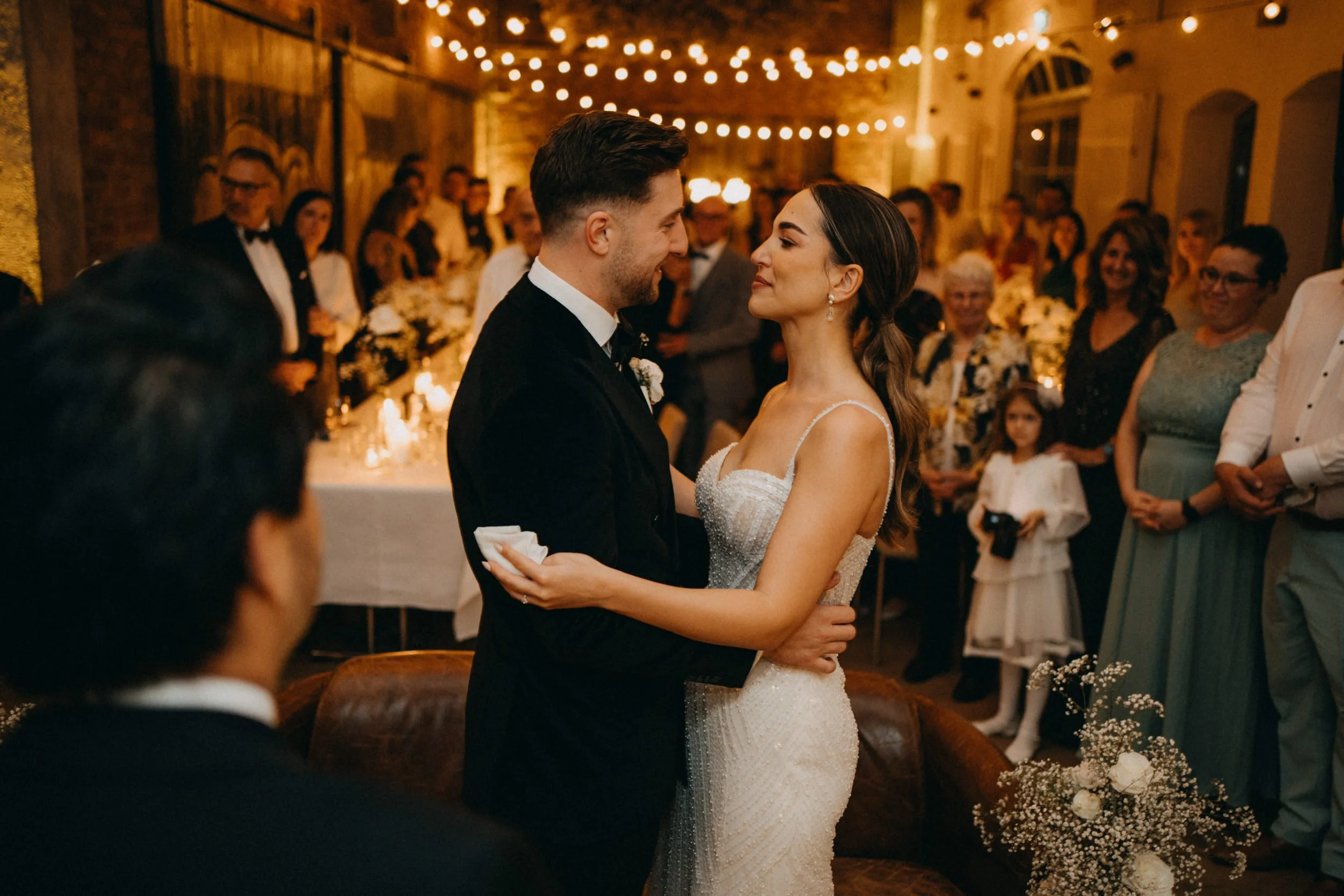 Ein Hochzeitspaar tanzt während ihrer Feier, umgeben von Gästen in einem rustikalen Raum mit warmem Licht und Dekorationen.
