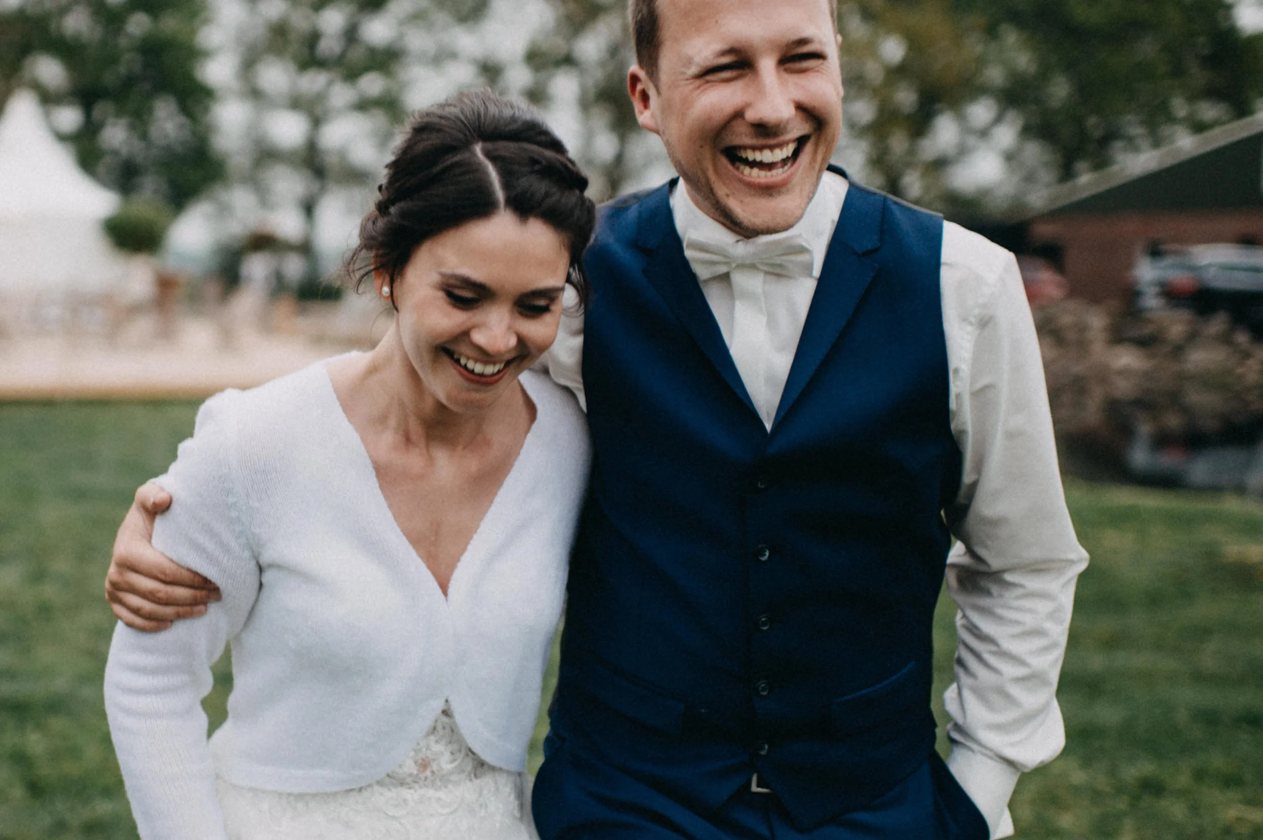 Ein Paar bei einer Hochzeit, das Arm in Arm lächelnd im Freien spazieren geht.