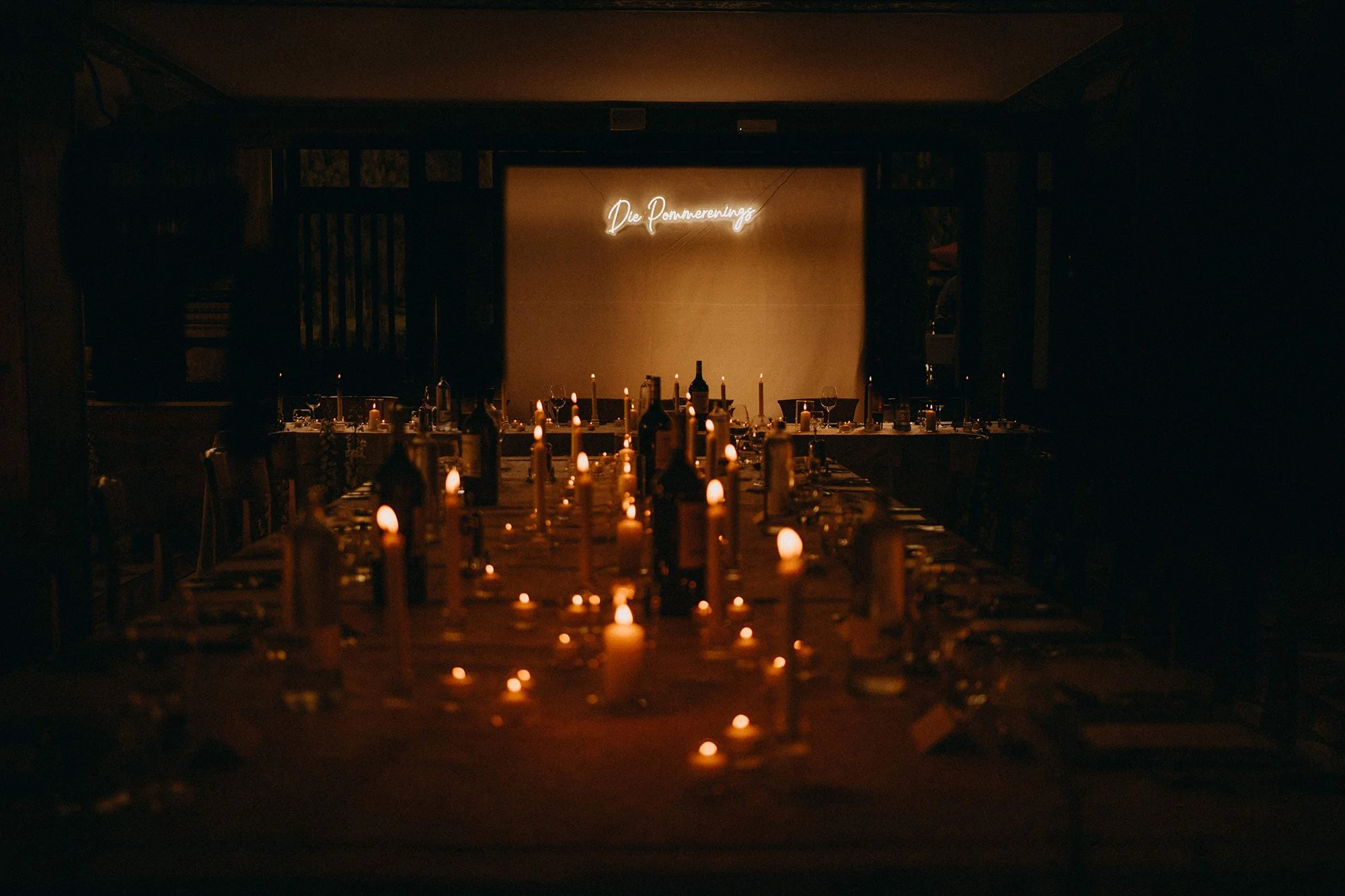 Ein dunkler Saal mit einem langen Esstisch, der mit Kerzen beleuchtet wird, und an der Wand hängt eine Neon-Schrift mit der Aufschrift 'Die Pommerners'.