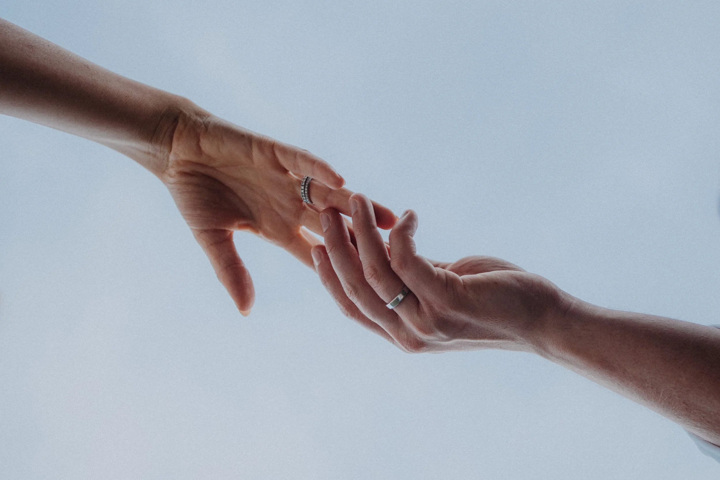 Zwei Hände, eine ältere mit Armband, die eine jüngere Hand berühren, vor hellem, neutralem Hintergrund.