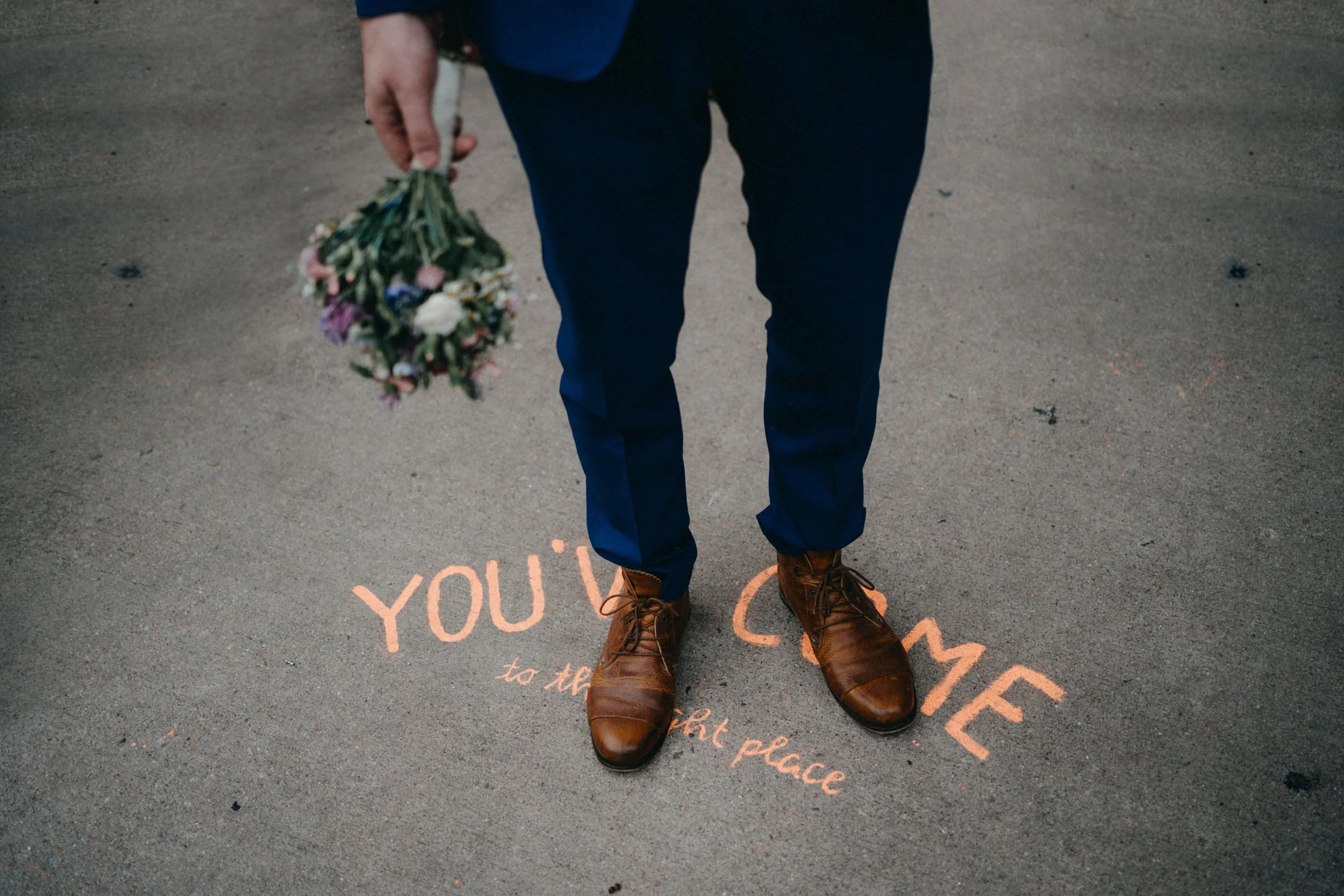 Eine Person mit braunen Stiefeln und dunklen Hosen steht auf einer Straße, die aufgemalt ist mit den Worten 'YOU' und 'to the'. Die Person hält einen Blumenstrauß in der Hand.