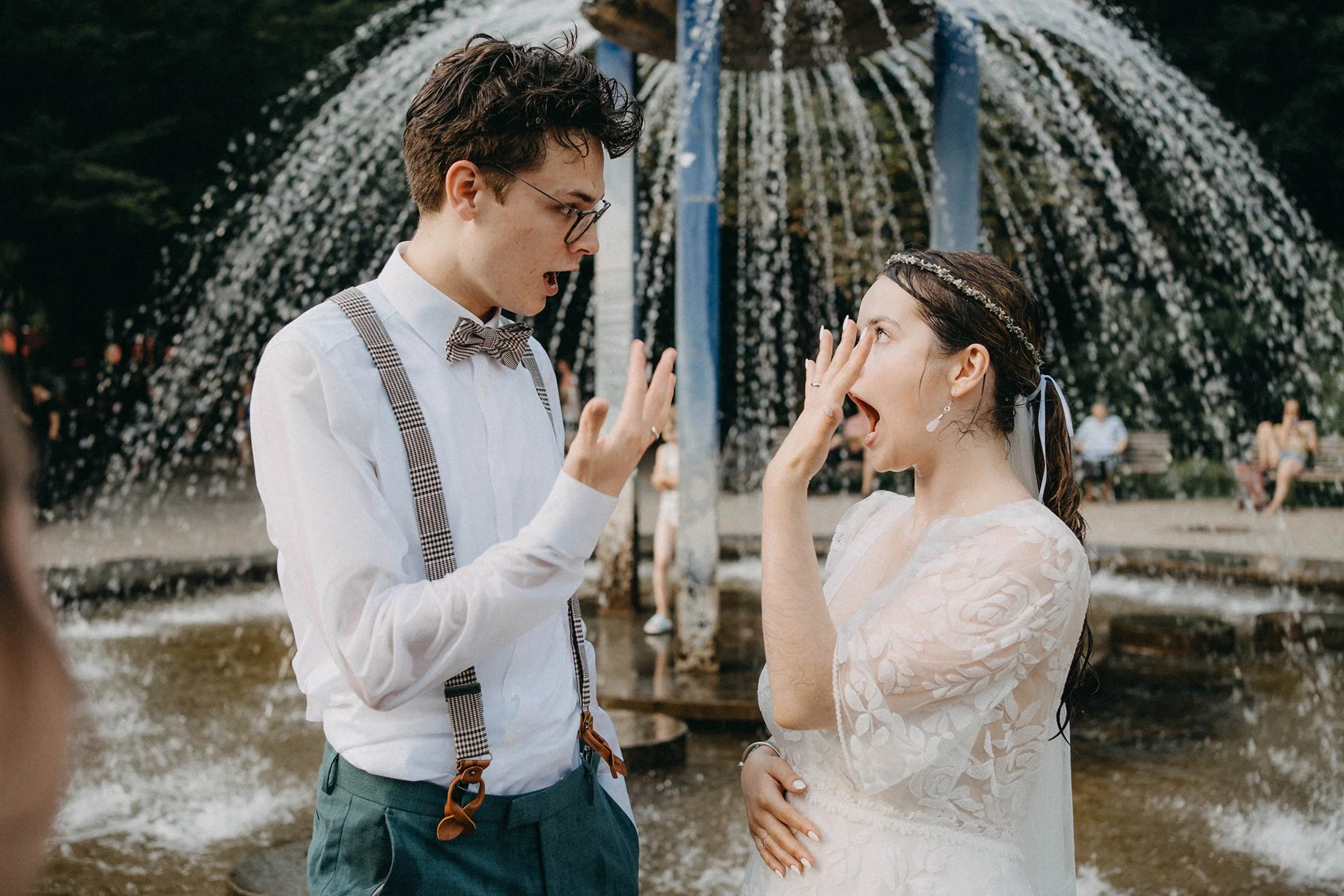 Ein junges Paar bei einer Hochzeit am Wasser, der Mann trägt eine weiße Hemd mit Hosenträgern und einer Fliege, die Frau trägt ein weißes Spitzenkleid mit einem Blumenkranz im Haar, sie sind im Gespräch und scheinen sich zu amüsieren.
