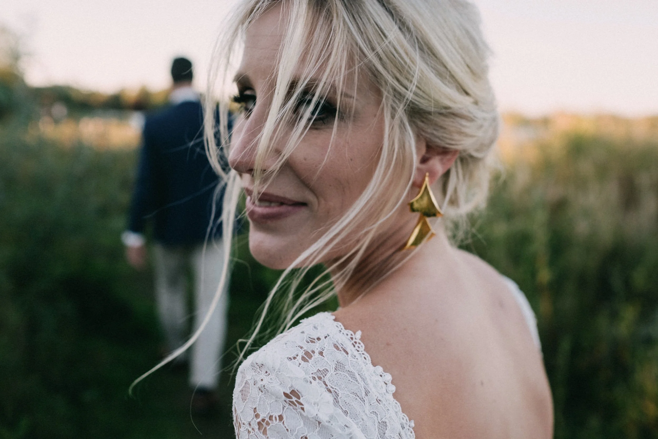 Nahaufnahme eines lächelnden blonde Frau in einem weißen Spitzenkleid mit großen goldenen Ohrringen, draußen bei Sonnenuntergang.