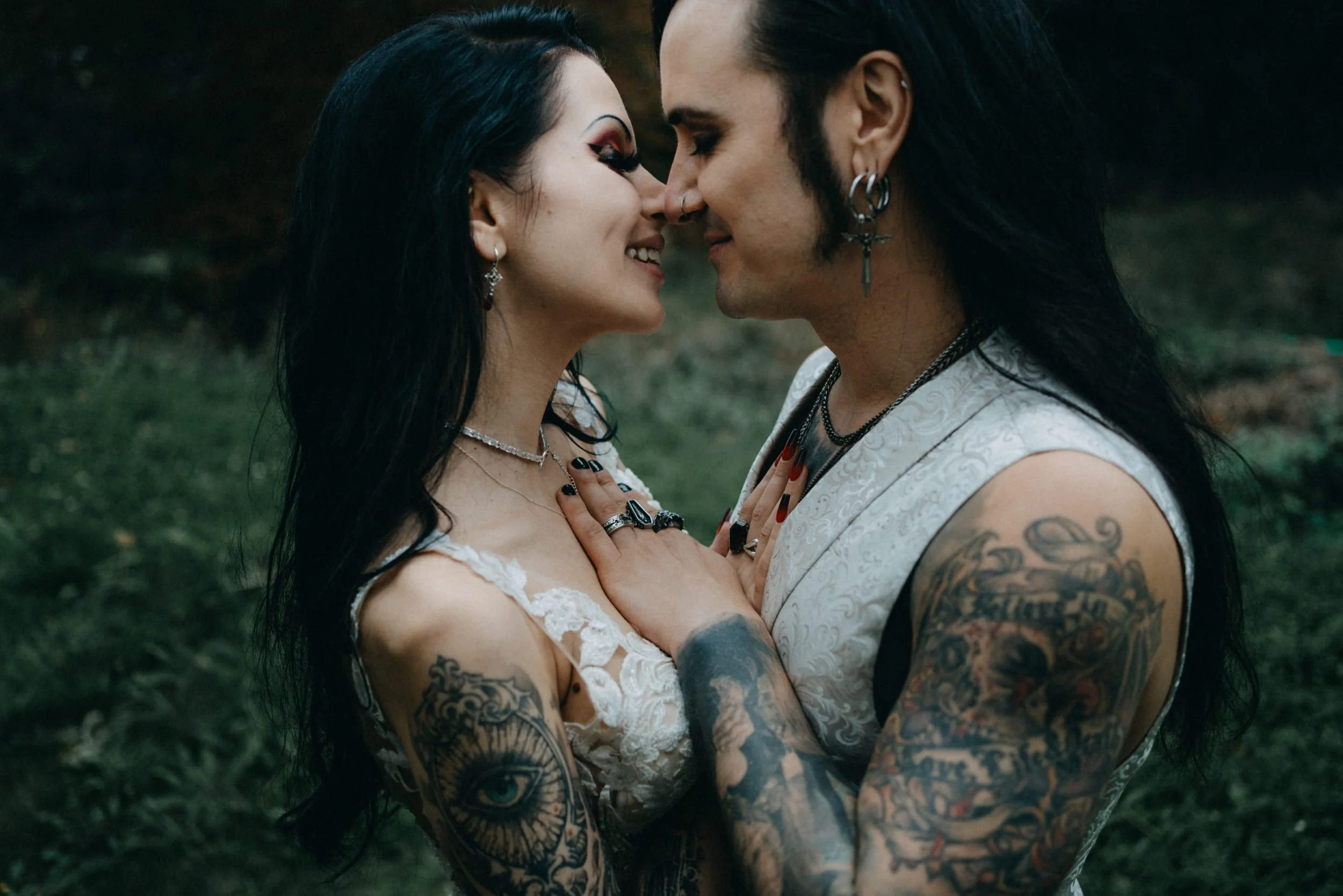 Zwei Menschen mit Tattoos und Piercings stehen sich Nah, berühren ihre Nasen und lächeln sich an, im Hintergrund grüner Wald.