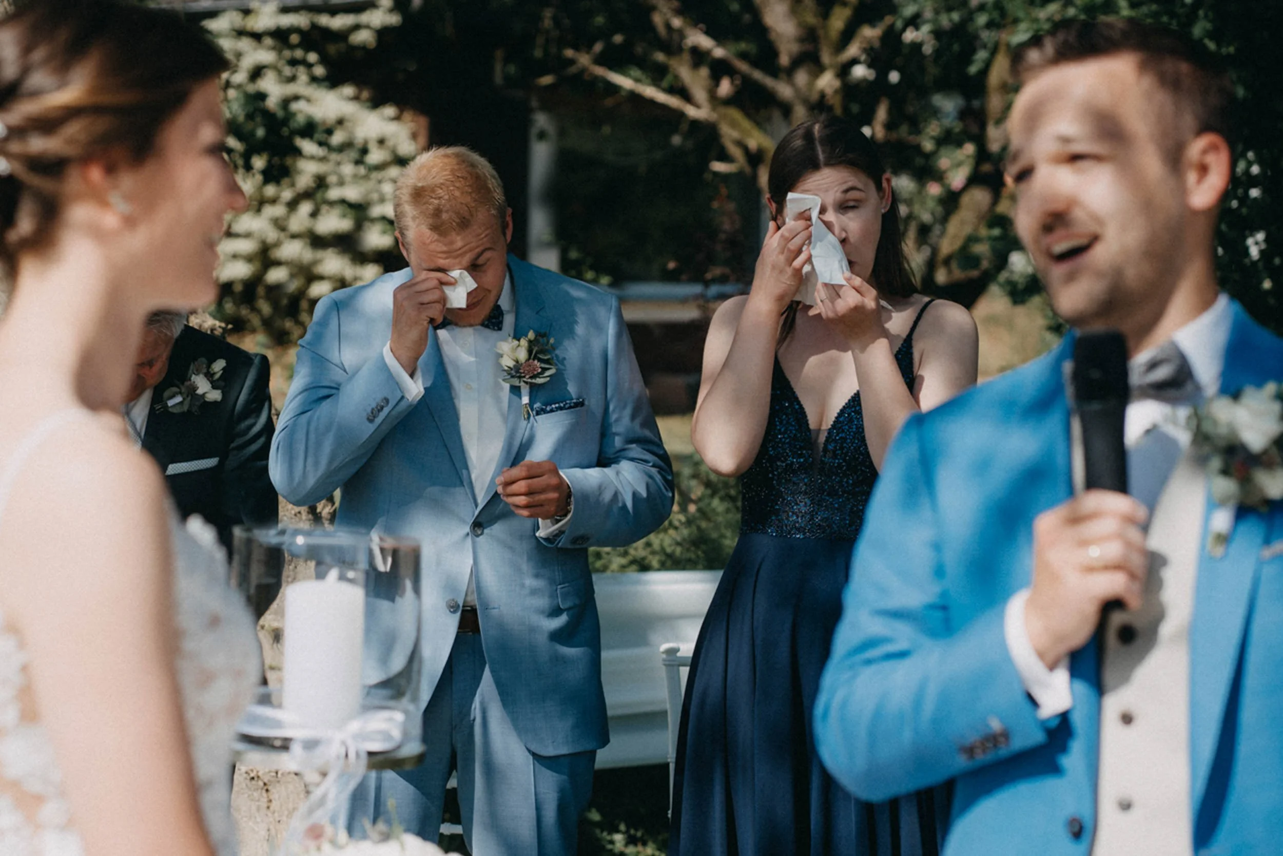 Hochzeitsgesellschaft tröstet sich bei Regen im Garten, einige zeigen emotionale Reaktionen, andere lachen.