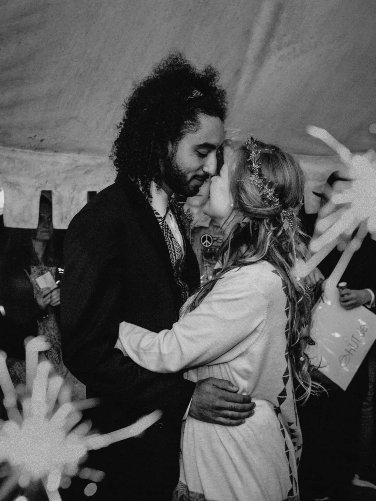 Ein Mann mit dunklem, lockigem Haar und Bart umarmt eine Frau mit langen, gewellten Haaren, die eine Blumenkrone trägt, bei einer Nähezeremonie, vermutlich einer Hochzeit, in einem Raum mit einer schwarzen und weißen Dekoration.