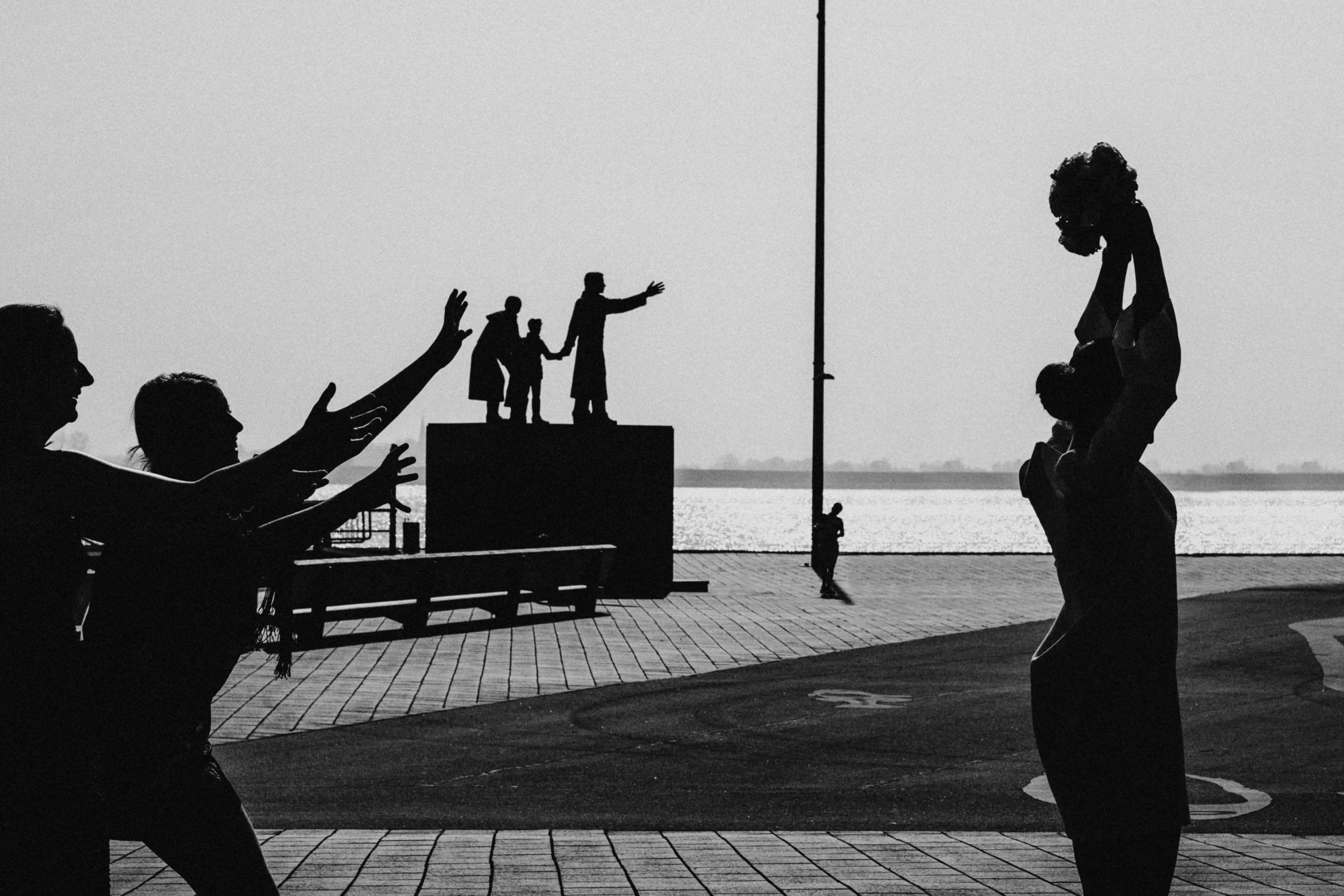 Silhouetten von Menschen, die entlang einer Uferpromenade spazieren, mit einem Paar, das Händchen hält, im Hintergrund. Eine Person trägt ein Baby, eine andere hat eine Hand zum Winken erhoben. Es ist wahrscheinlich Abend oder Sonnenuntergang. Im Hin