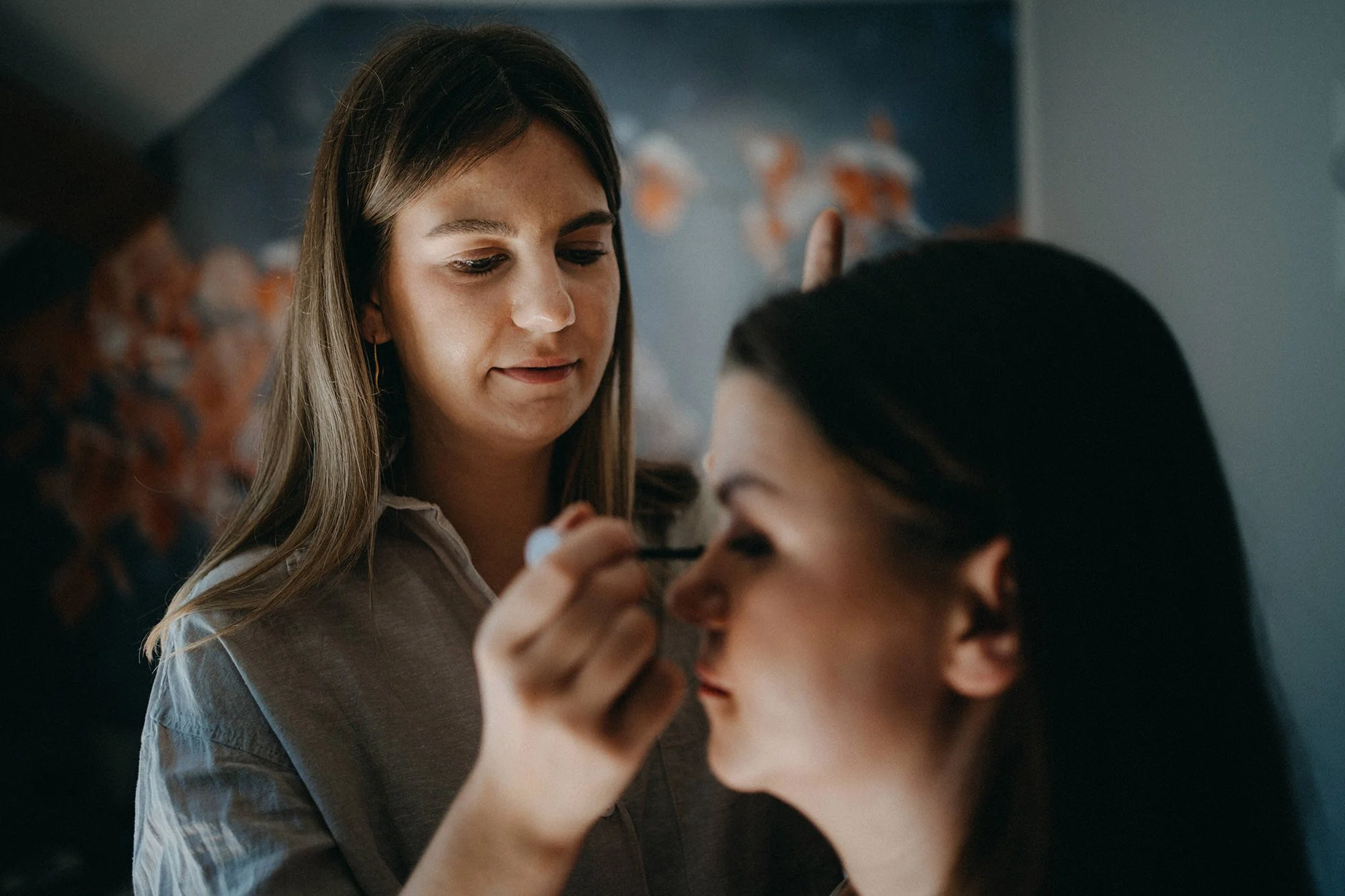 Zwei Frauen, die eine Make-up Anwendung durchführen, eine mit braunen Haaren, die andere mit schwarzen Haaren. Im Hintergrund ist eine dunkle Wand mit einem bunten Muster.