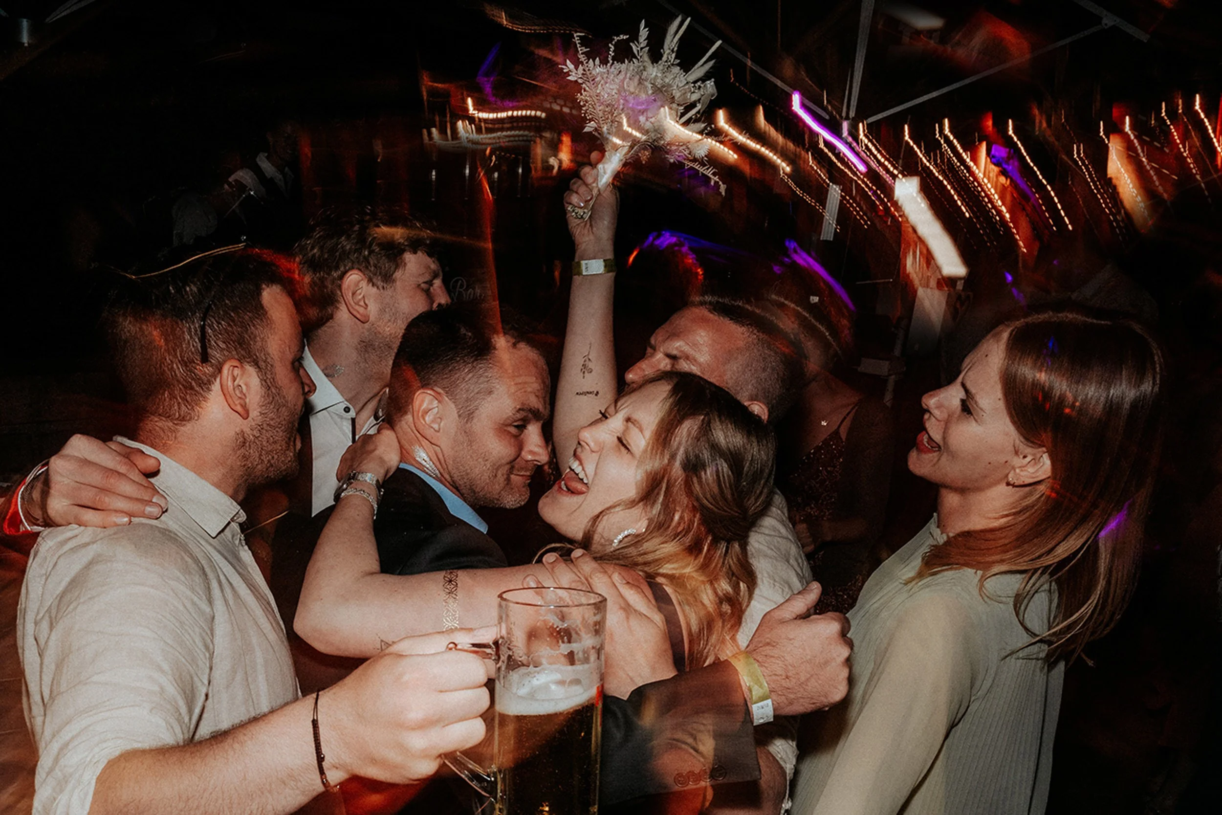 Menschen feiern, umarmen sich, trinken Bier, lachen, umgeben von buntem Licht. Es scheint eine festliche Party zu sein.