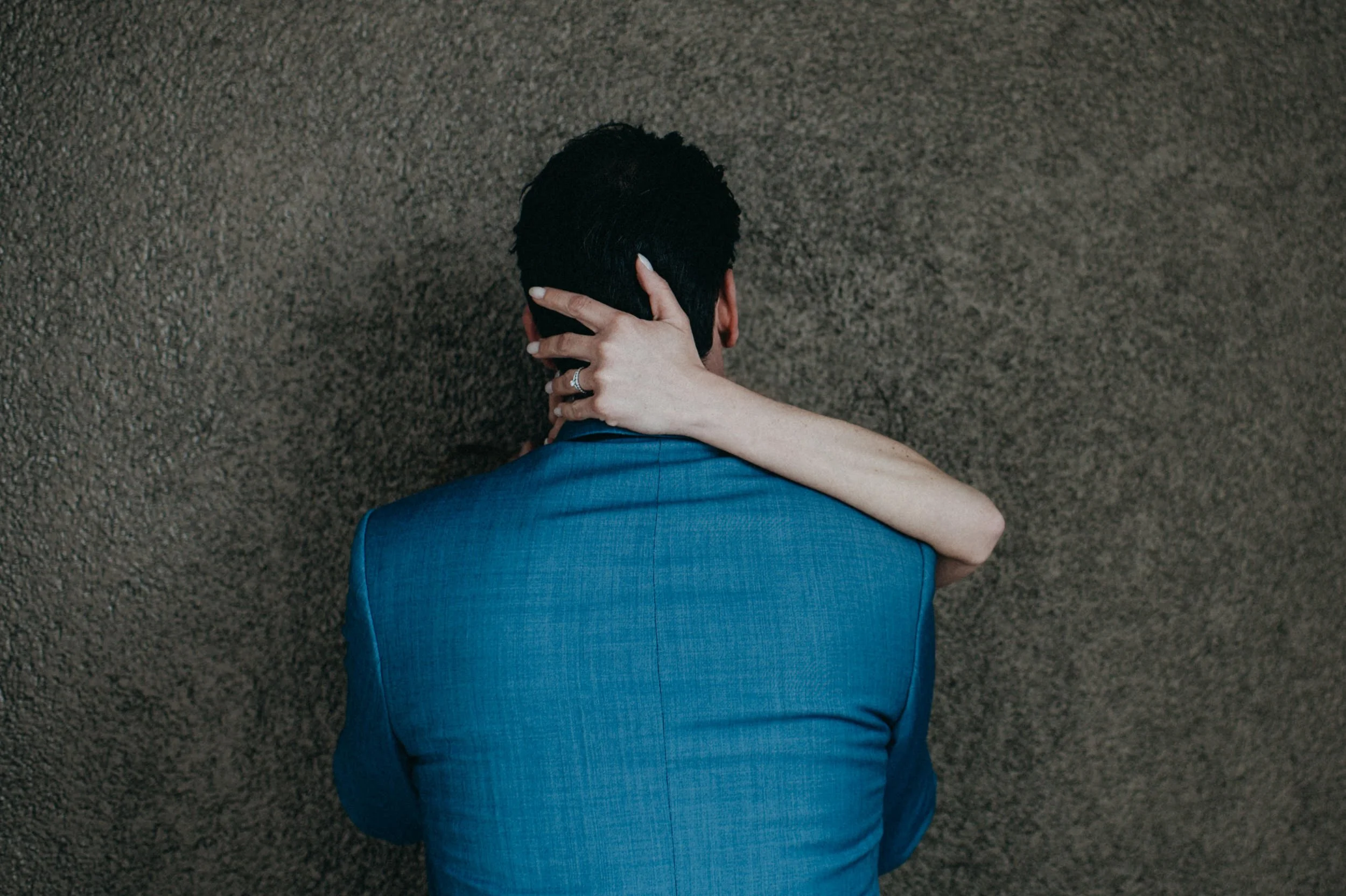 Eine Person in einem blauen Anzug sitzt am Boden, mit dem Rücken zur Kamera, auf dunklem Teppichboden, die rechte Hand hält den Nacken.