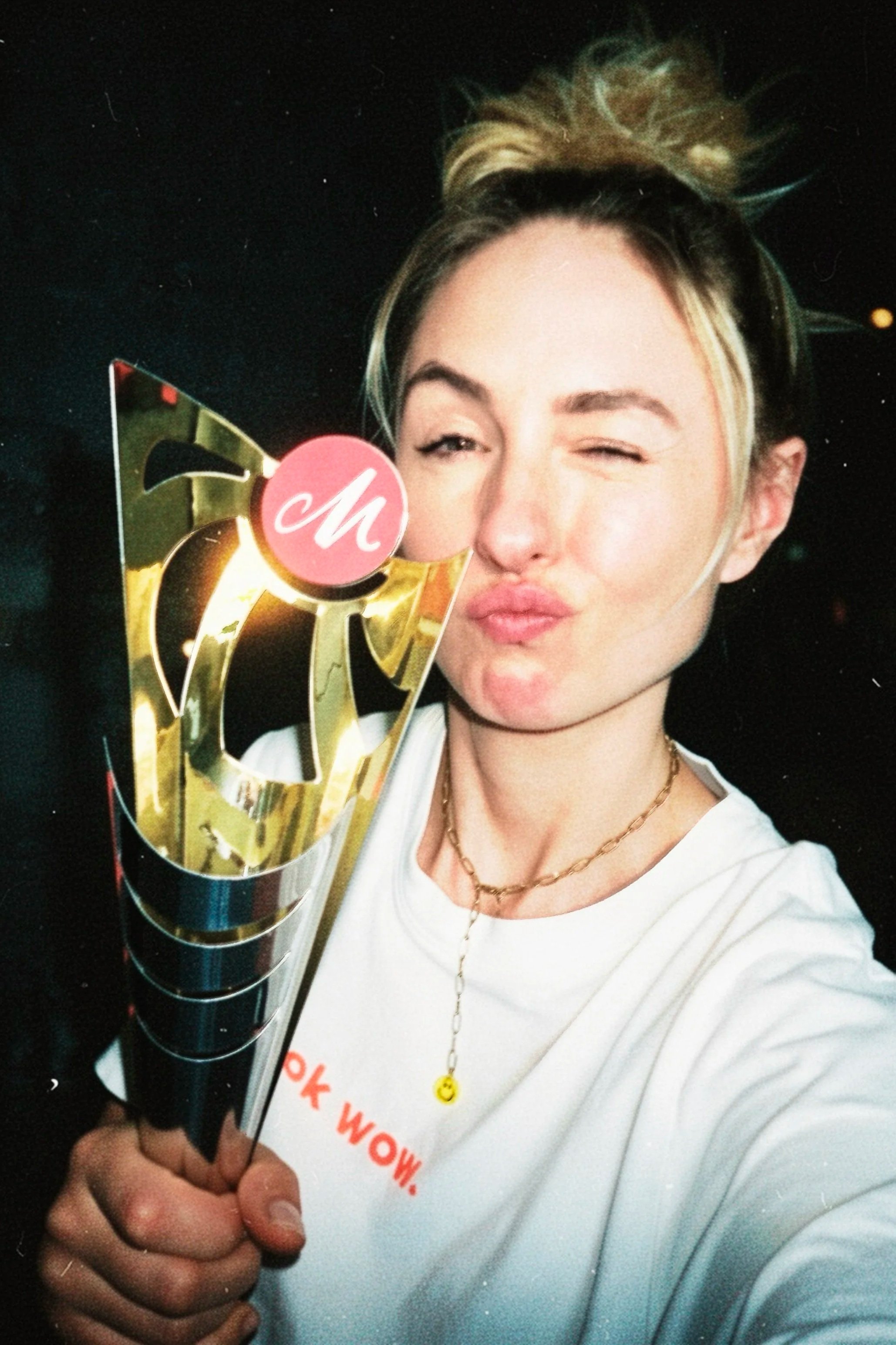 Junge Frau, die eine Trophäe hält und in die Kamera lächelt, mit einem zarten Goldhalskette und einem weißen T-Shirt mit roten Schriftzug. Sie winkt mit der Hand und trägt einen lockeren Dutt hoch oben auf dem Kopf. Der Hintergrund ist dunkel und nur einige kleine Lichtpunkte sind sichtbar.