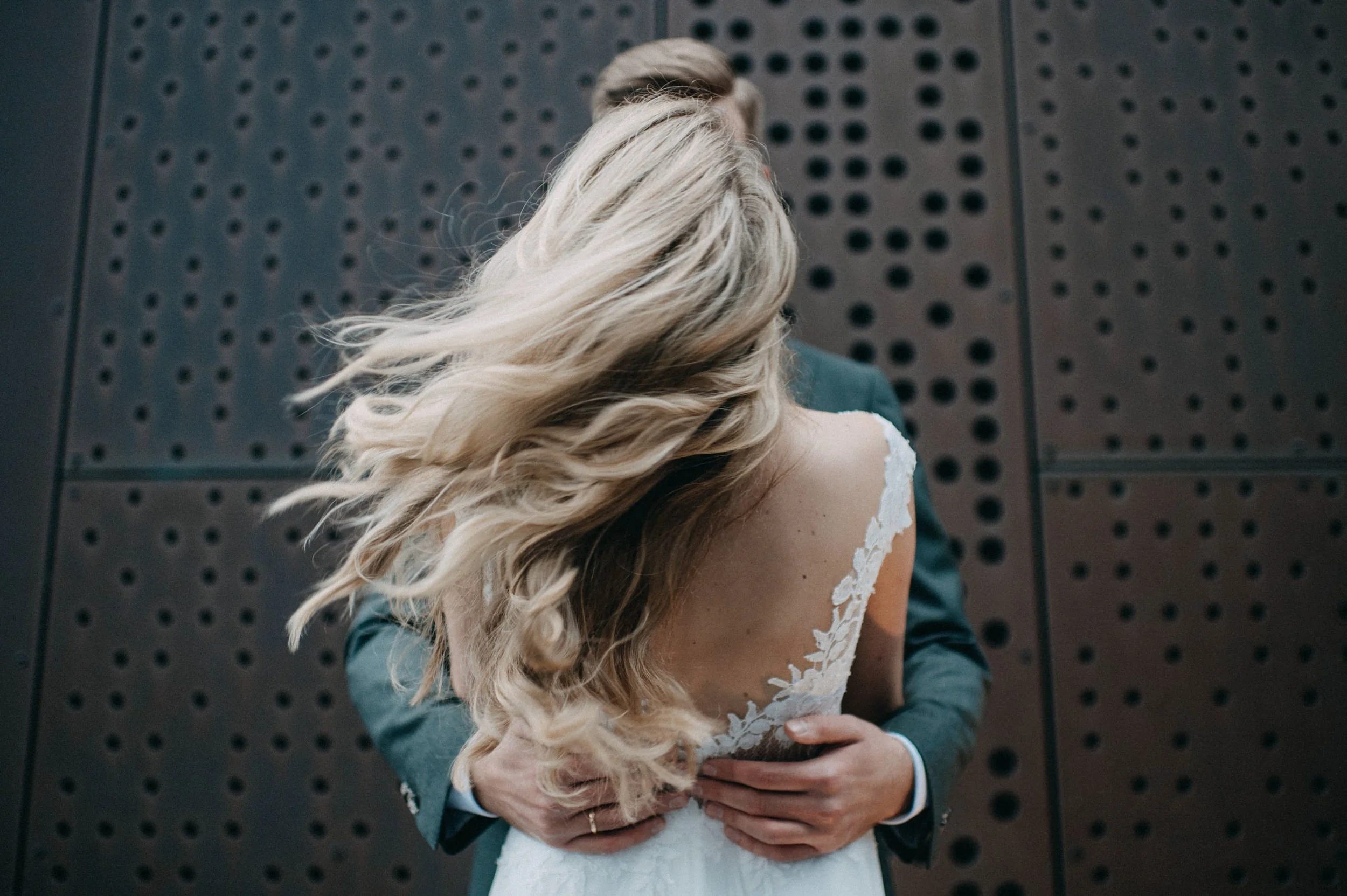 Ein Brautpaar umarmt sich vor einer dunklen, gelochten Metallwand. Das Brautkleid hat weiße Spitze, die Braut hat lange, blonde, lockige Haare, der Bräutigam trägt einen dunklen Anzug.