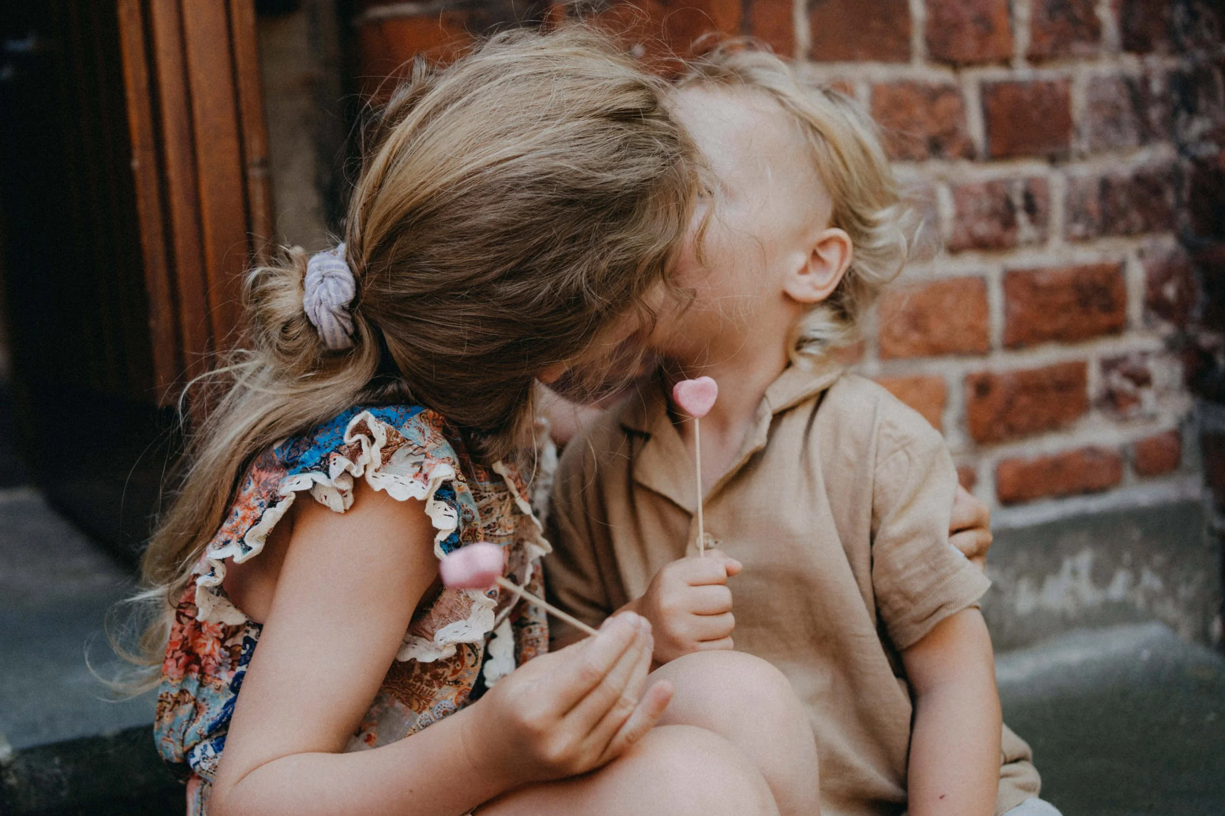 Zwei Kinder, ein Mädchen mit braunen Haaren und einer Schleife, ein Junge mit blonden, lockigen Haaren, sitzen eng zusammen vor einer roten Backsteinmauer und teilen sich eine Lutscher.