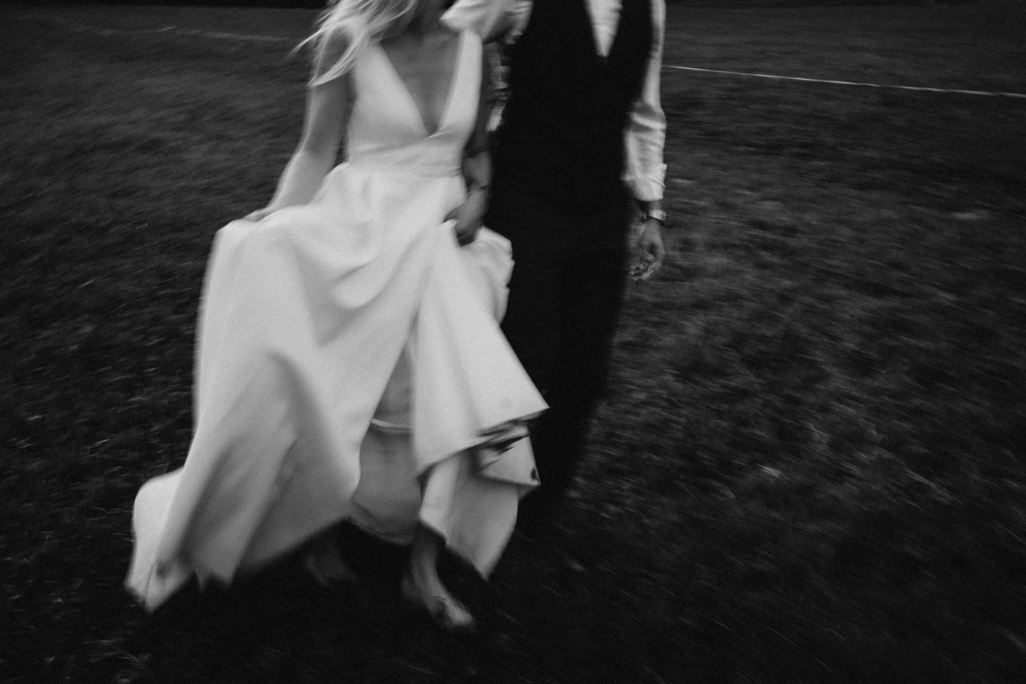 Schwarz-weiße Aufnahme eines Paares, das Hand in Hand läuft. Die Frau trägt ein elegantes Kleid mit weitem Saum, der beim Laufen schwingt, der Mann trägt formale Kleidung. Der Hintergrund ist unscharf, die Szene wirkt dynamisch und romantisch.