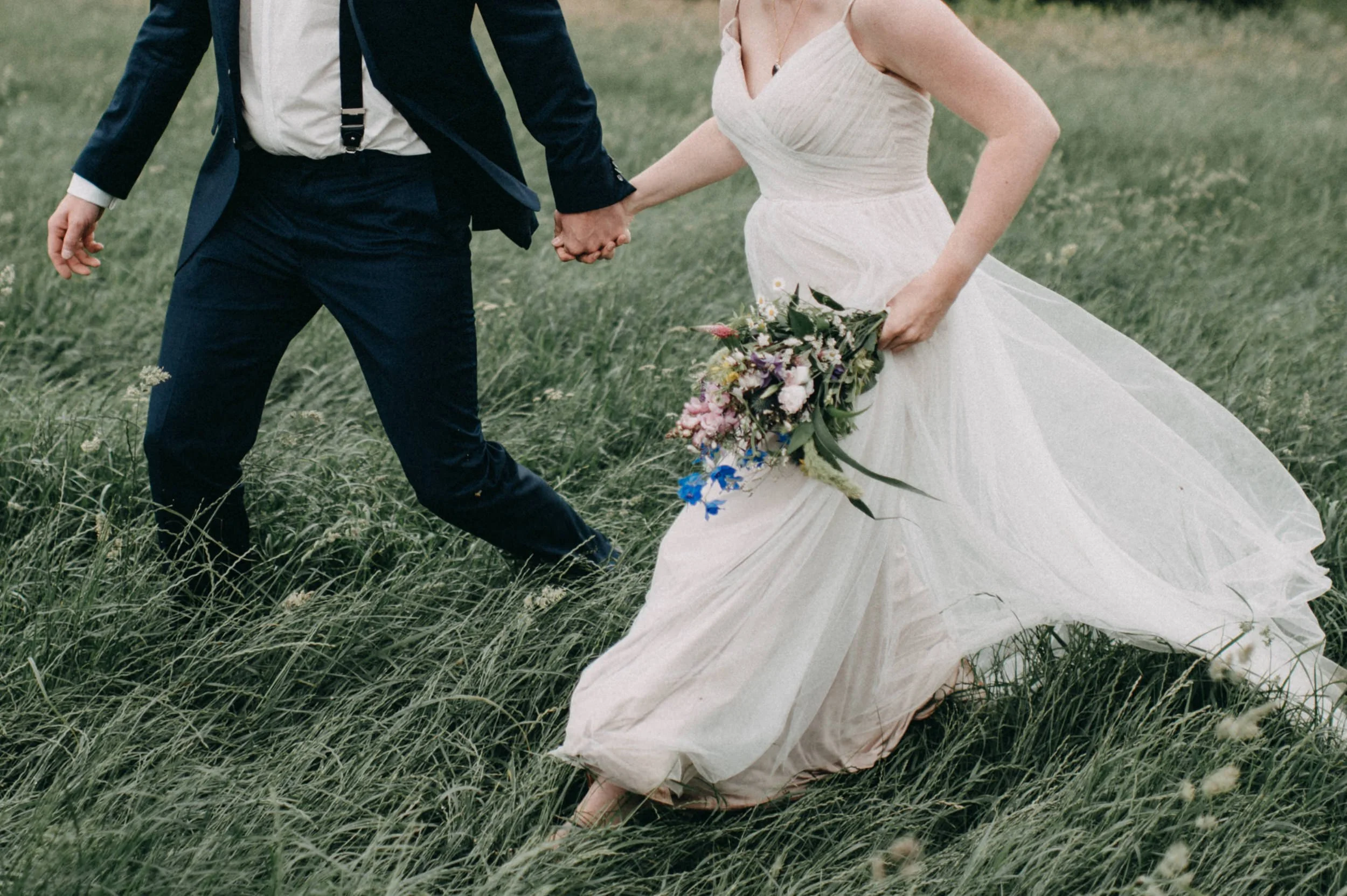 Ein Brautpaar hält Händchen in einem Feld, die Frau trägt ein weißes Hochzeitskleid mit Blumenstrauß, der Mann trägt einen dunklen Anzug.