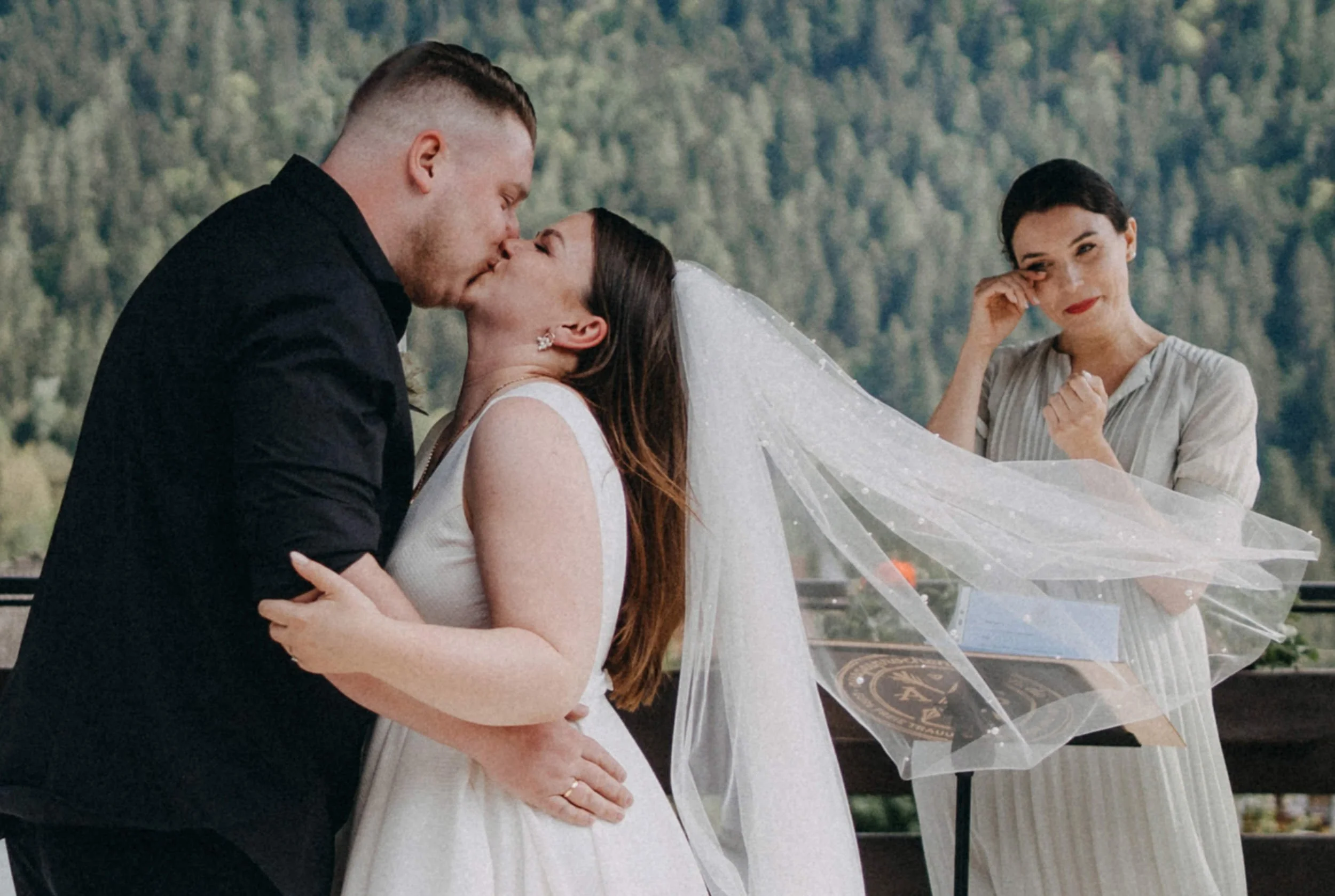 Ein Brautpaar küsst sich bei ihrer Hochzeit, während eine Brautmutter neben ihnen lächelt und Tränen der Freude in den Augen hat.