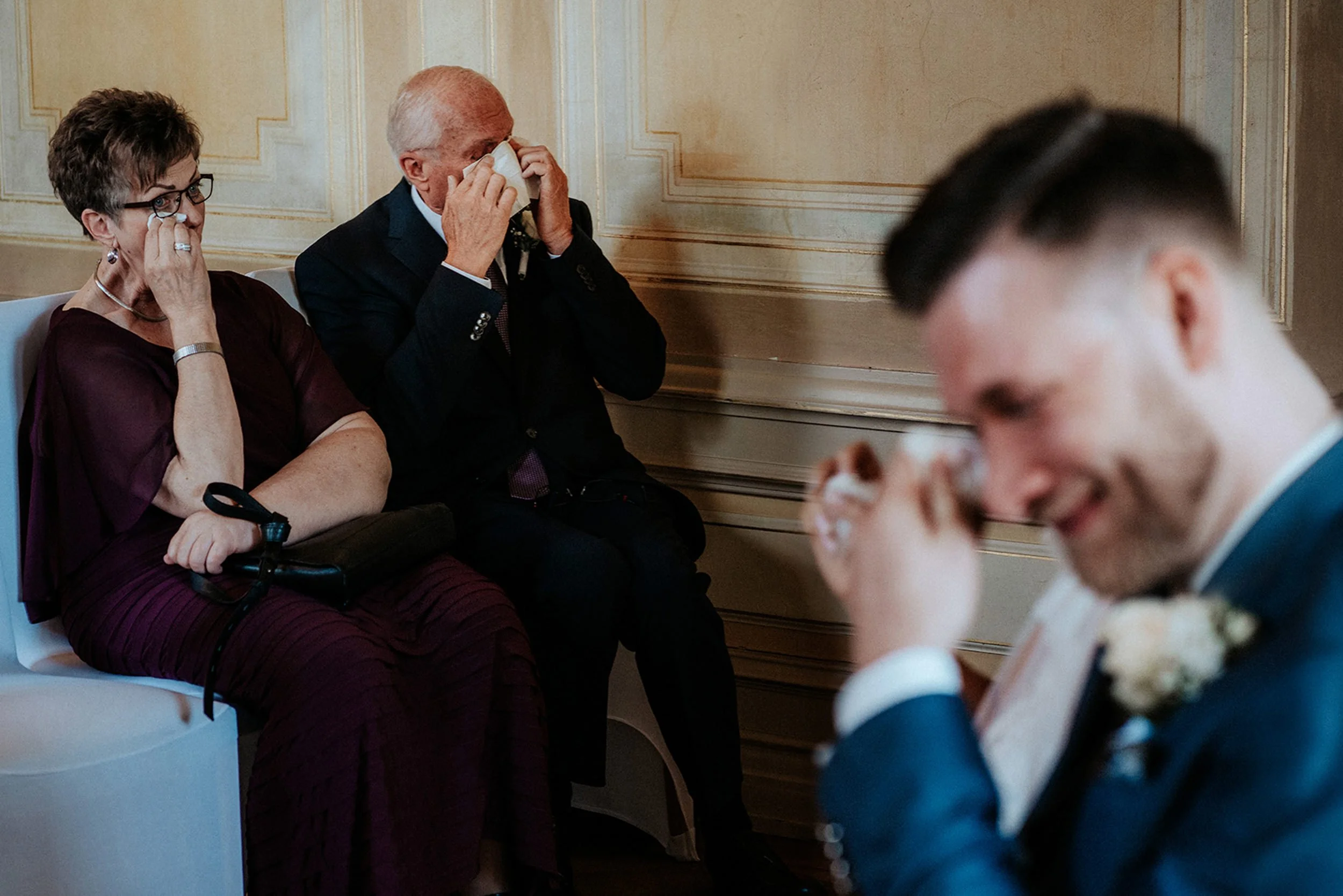 Älterer Mann und ältere Frau sitzen im Theater, die Frau hat eine Brille, trägt ein dunkelrotes Kleid und hat Tränen im Auge, während der Mann eine Tasse hält und an die Augen fasst.