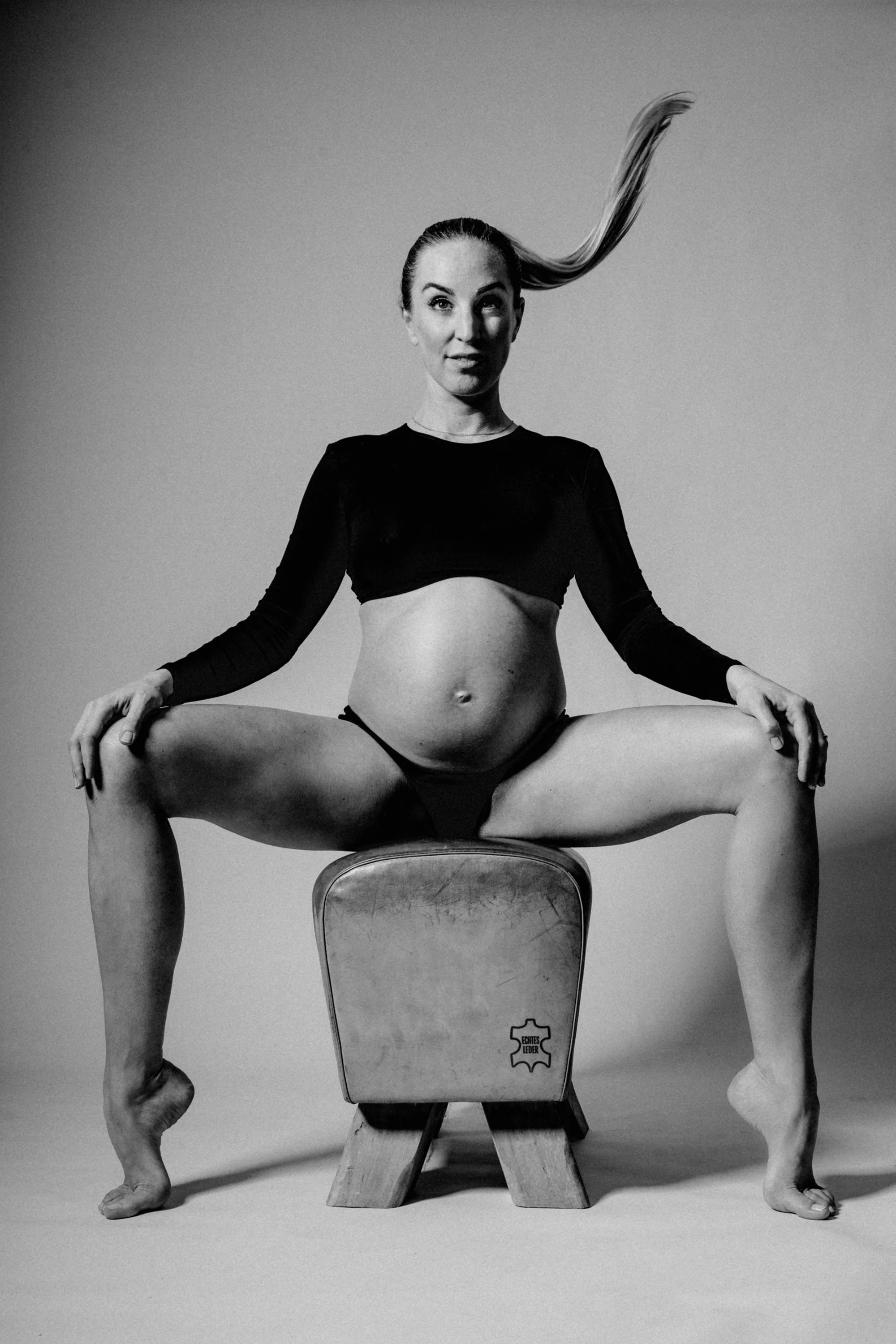 Schwarz-weiß-Foto einer Frau, die auf einem Vintage-Stuhl im Schneidersitz sitzt, mit den Armen auf den Knien, in aufrechter Haltung, Haare nach oben hochgebunden, trägt ein schwarzes Langarmtop, und hat die Beine weit gespreizt.