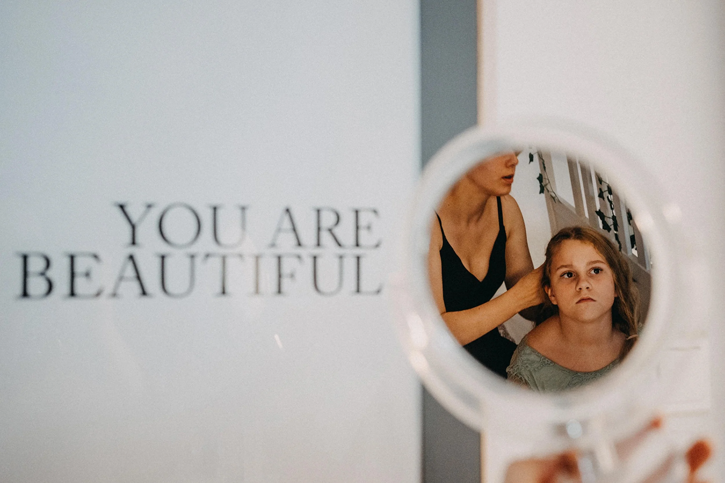 Blick in einen Spiegel, der von einem weißen Rahmen umgeben ist, bei dem eine Mutter und ein Kind zu sehen sind. Auch ein Teil eines Posters mit dem Text "You are beautiful" ist sichtbar.