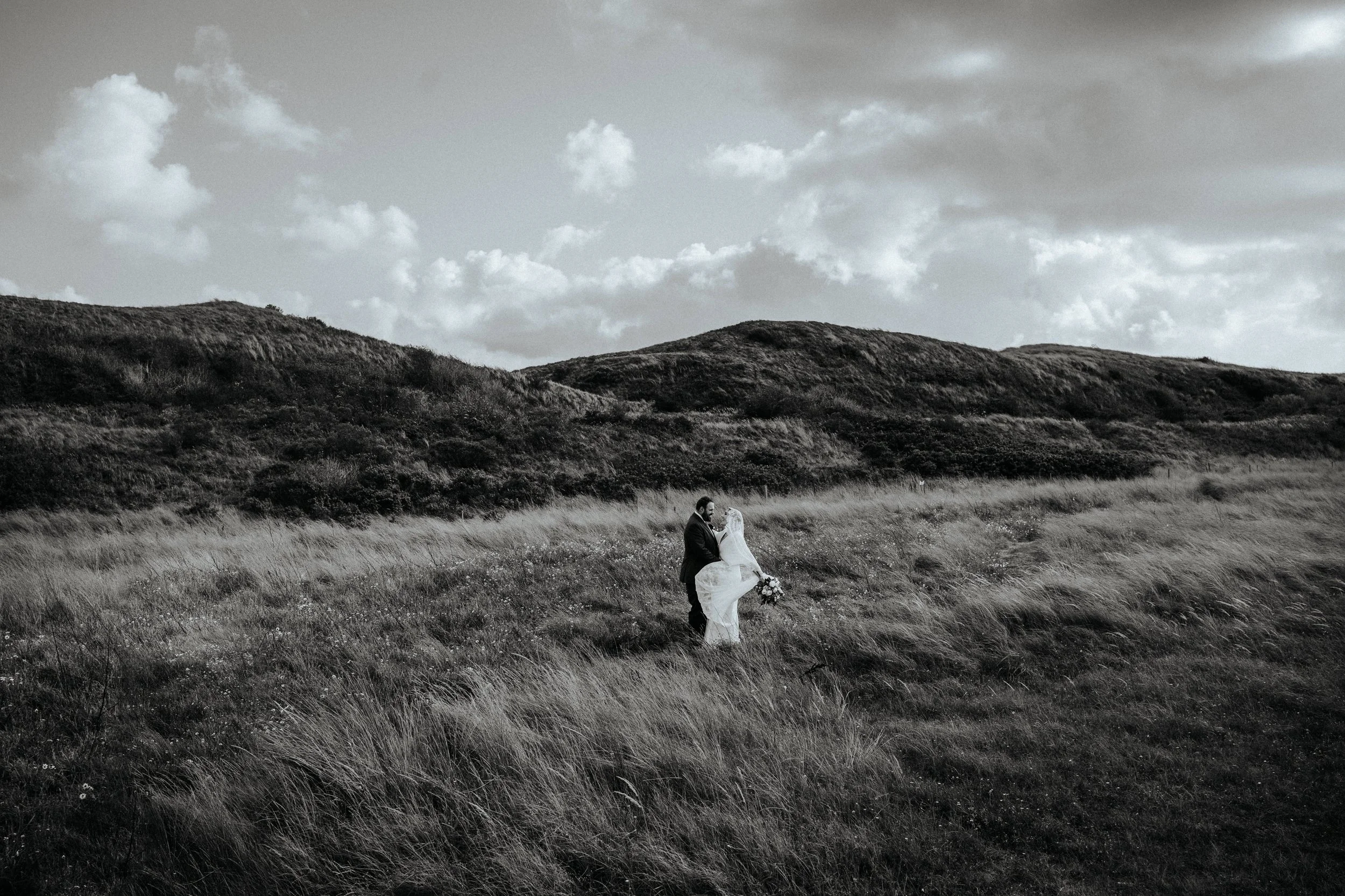 Ein Hochzeitspaar steht in einer offenen Wiese mit Bergen im Hintergrund und Wolken am Himmel, schwarz-weiß Foto.