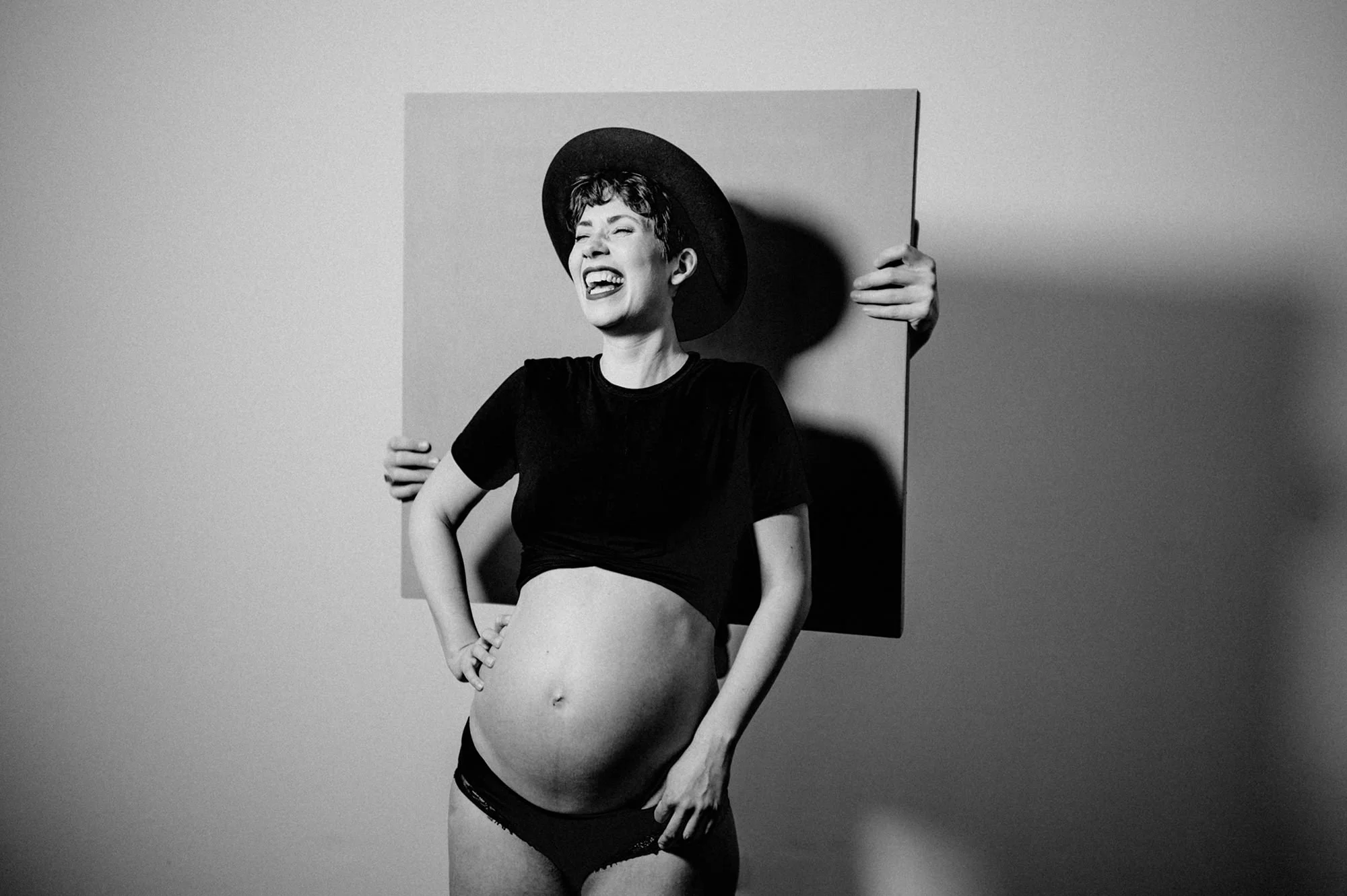 Schwarz-Weiß-Foto einer lachenden Frau in Unterwäsche, trägt einen Hut und steht vor einem quadratischen Hintergrund.
