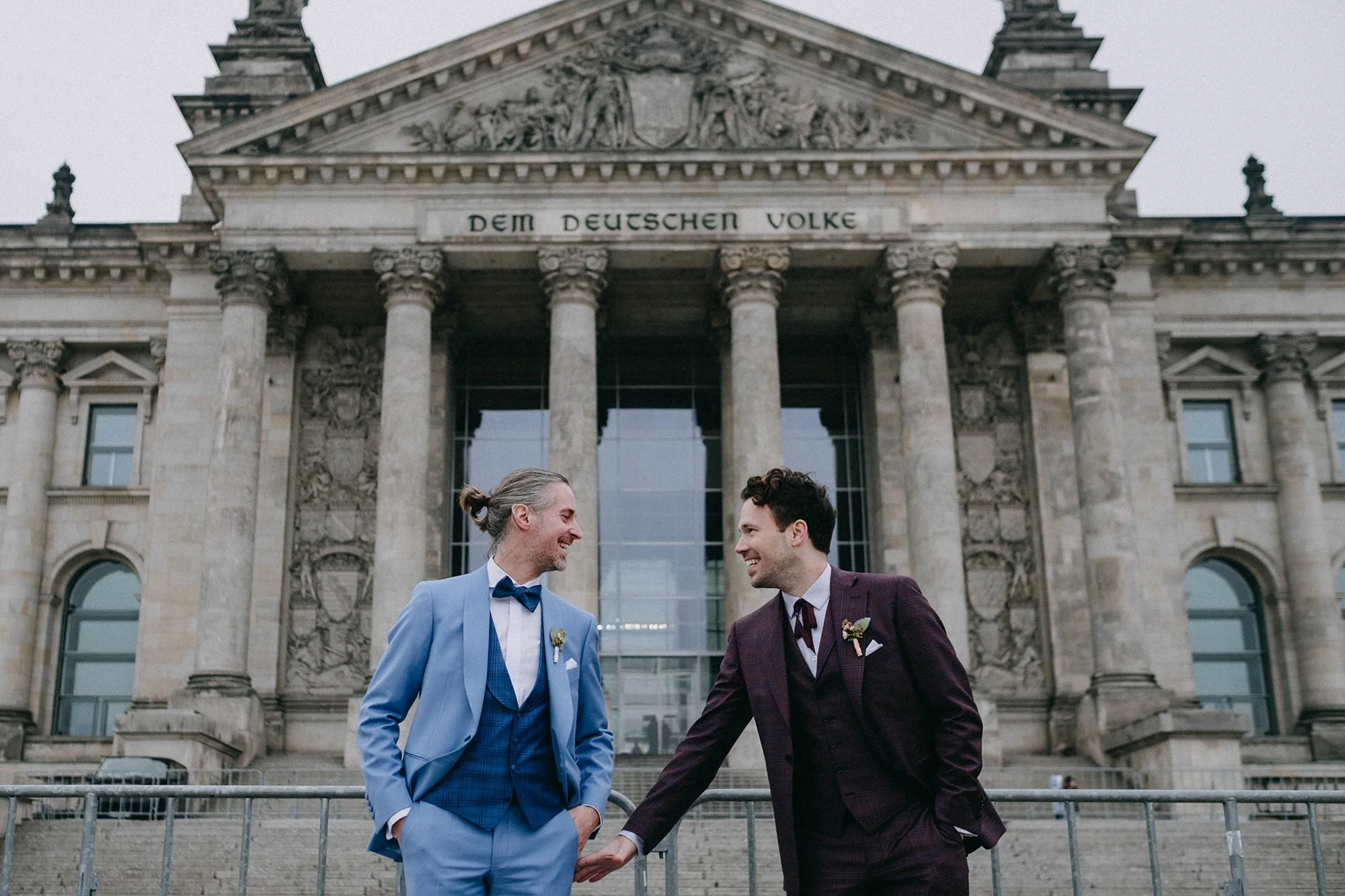 Zwei Männer in schicken Anzügen, die vor einem historischen Gebäude mit Säulen, Statuen und einer Inschrift stehen, lachen und schütteln sich die Hände.
