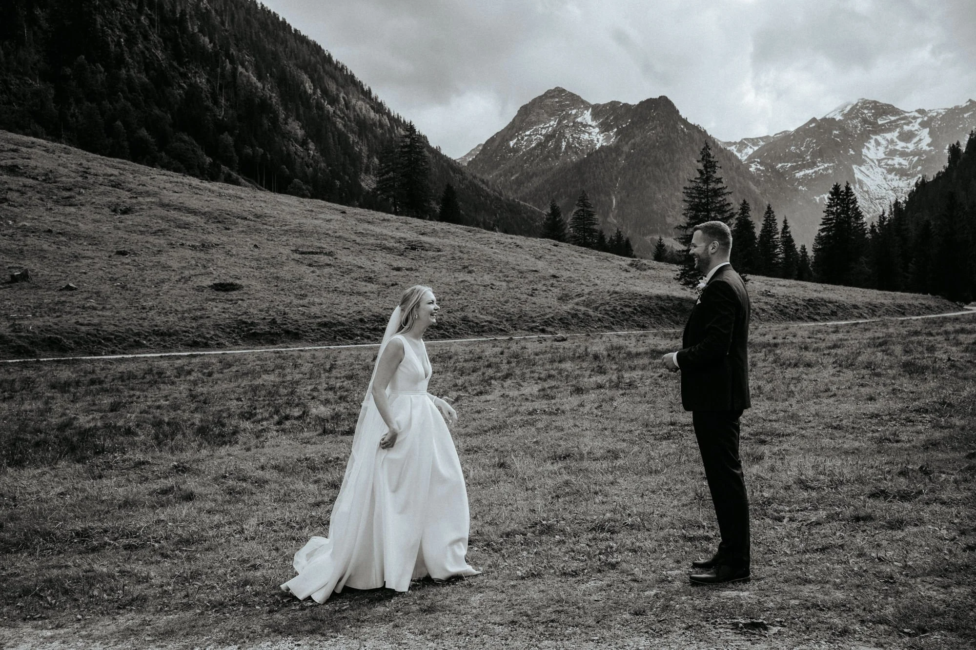 Ein Brautpaar steht sich in einer Berglandschaft gegenüber. Die Braut trägt ein langes weißes Kleid und ein Schleier, der Bräutigam trägt einen dunklen Anzug. Die Umgebung ist von Bergen mit Schneefeldern und Tannenbäumen geprägt. Das Bild ist in Schwarz-Weiß gehalten.