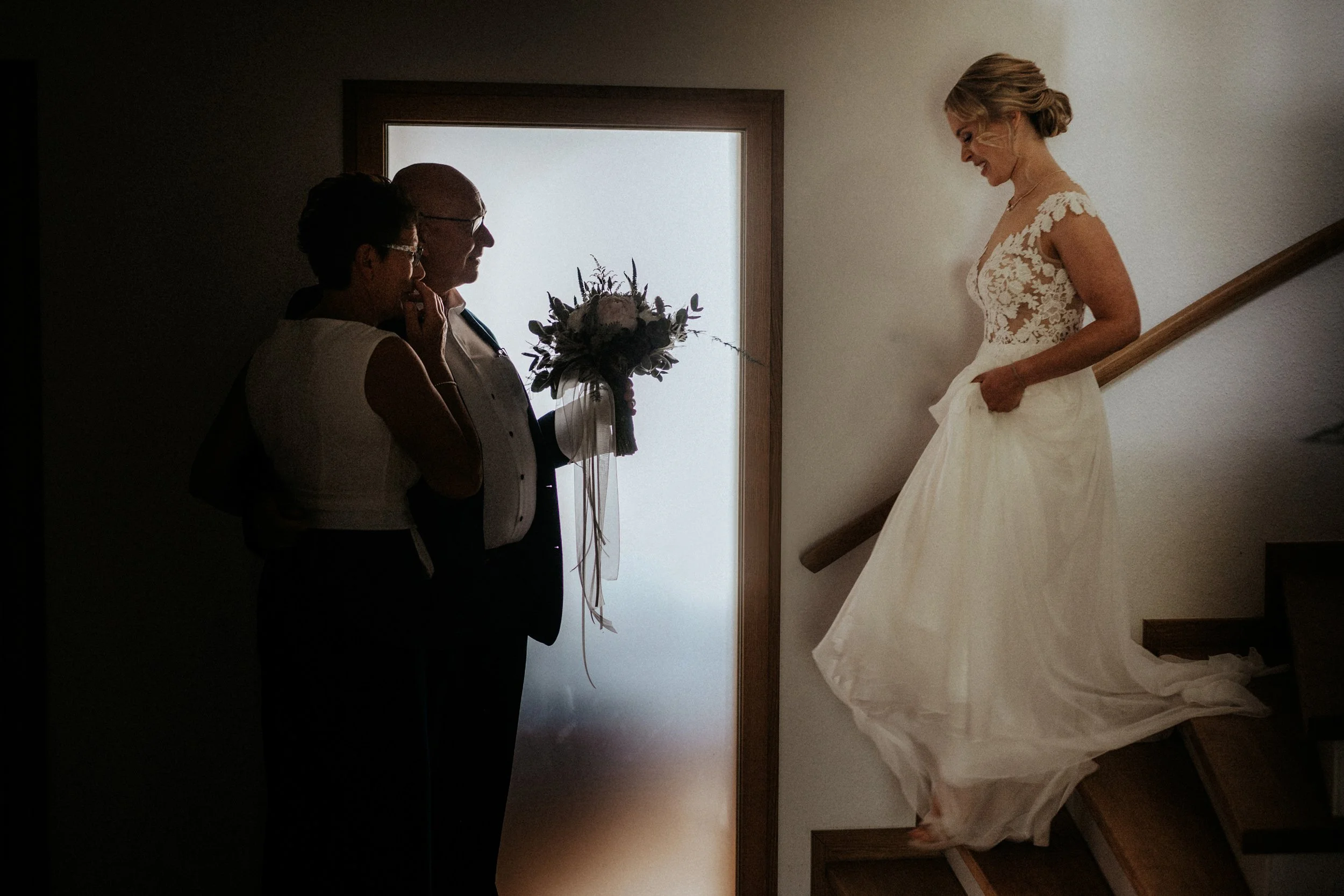 Eine Hochzeitsszene mit einer Braut in einem weißen Kleid, die auf einer Treppe steht, und drei Personen im Hintergrund, die miteinander sprechen.