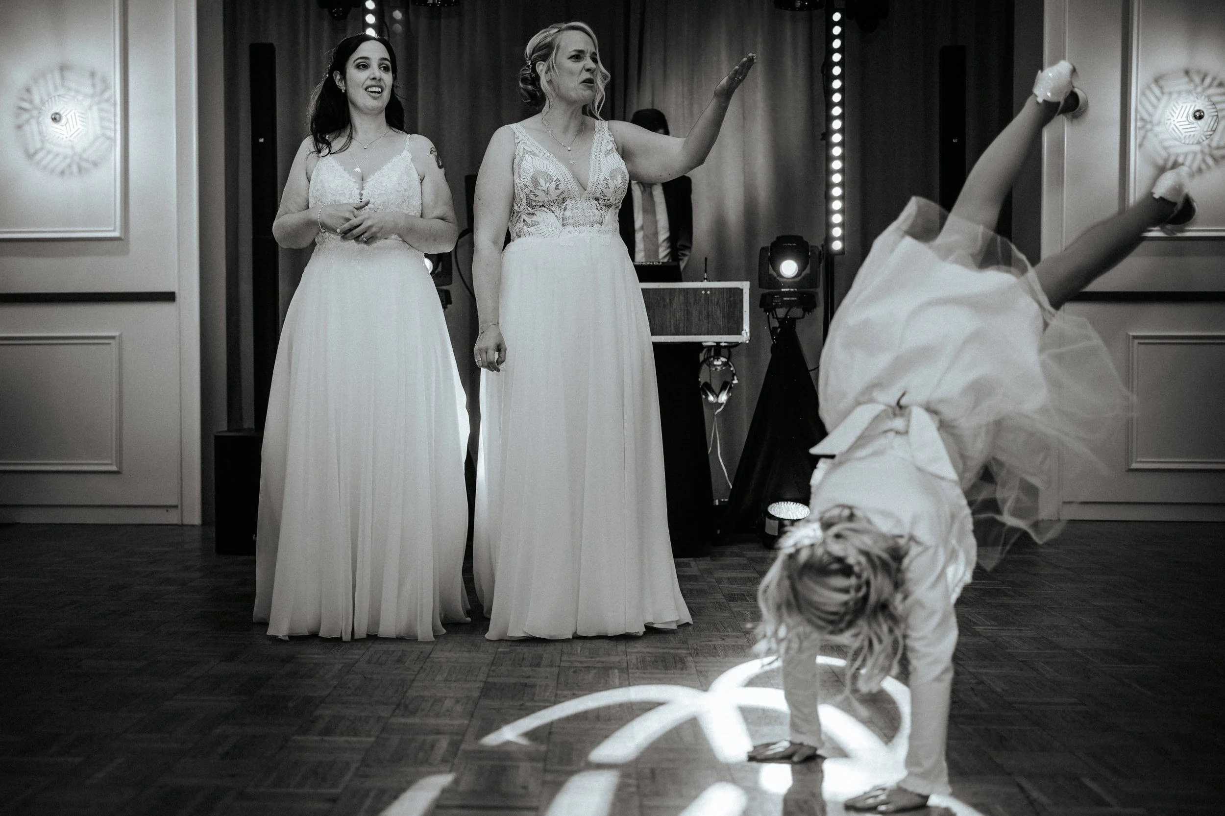 Zwei Frauen in weißen Brautkleidern und ein Mädchen in Tutu-Tanzkostüm im schwarzen und weißen Foto bei einer Hochzeit, die auf der Tanzfläche tanzen.
