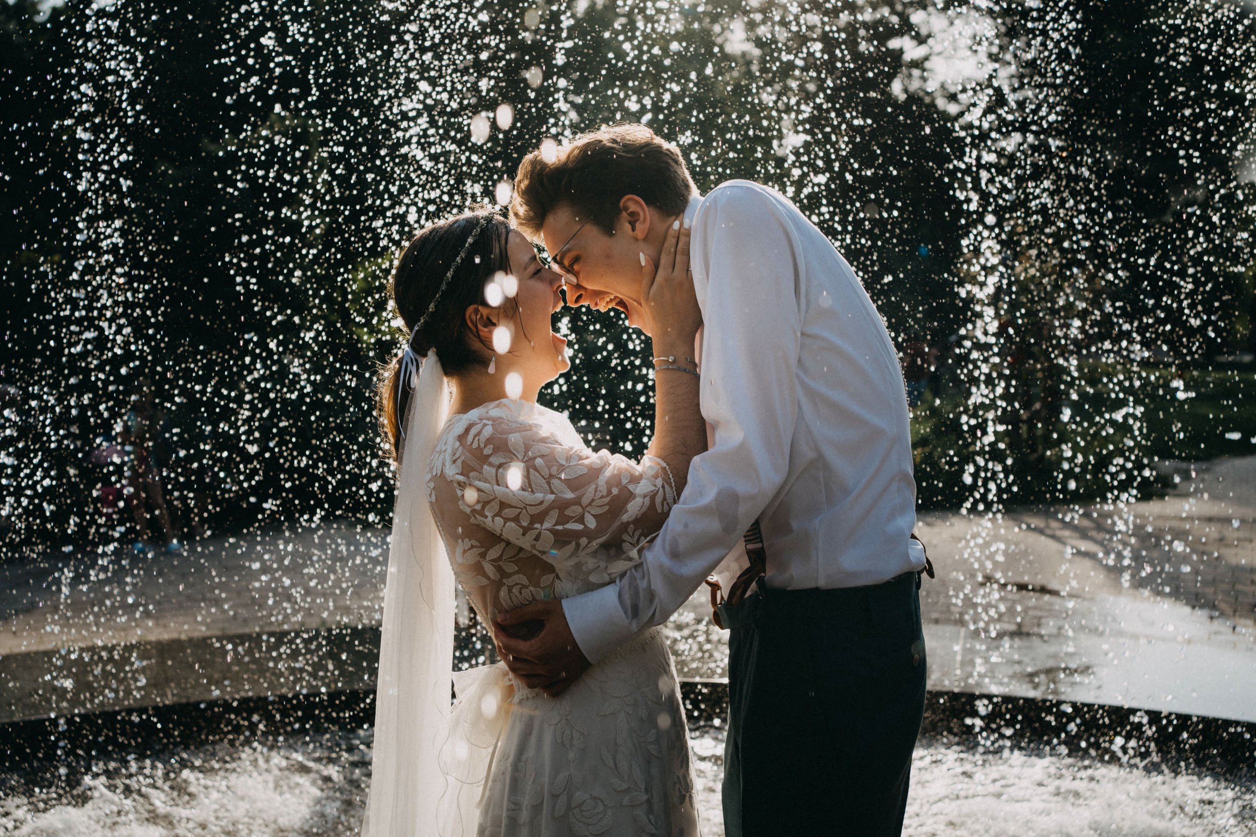 Ein Paar im Hochzeitkleidungsstück küsst sich unter einem Wasserregenguss im Freien, mit Sonnenlicht im Hintergrund.
