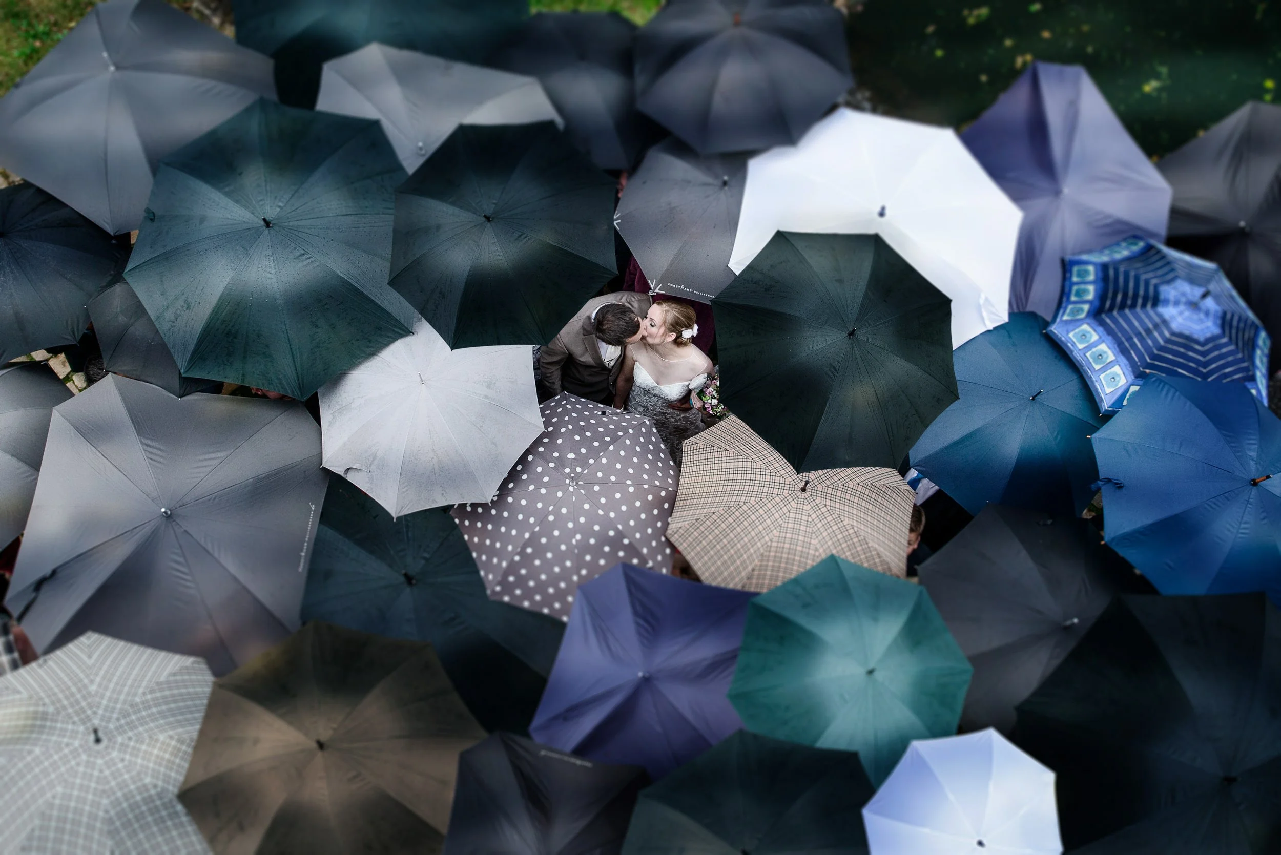 Zwei Menschen küssen sich unter einem Schirmdach aus Regenschirmen in verschiedenen Farben und Mustern bei Regen.