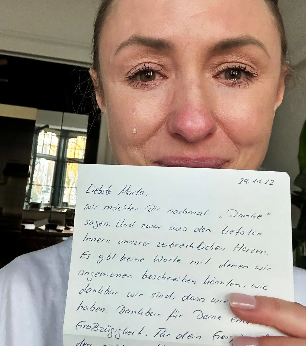 Eine Frau hält einen handgeschriebenen Brief vor das Gesicht, bei dem nur die Augen und die obere Gesichtshälfte sichtbar sind. Sie wirkt emotional, mit einem Tränentropfen an ihrer Wange.