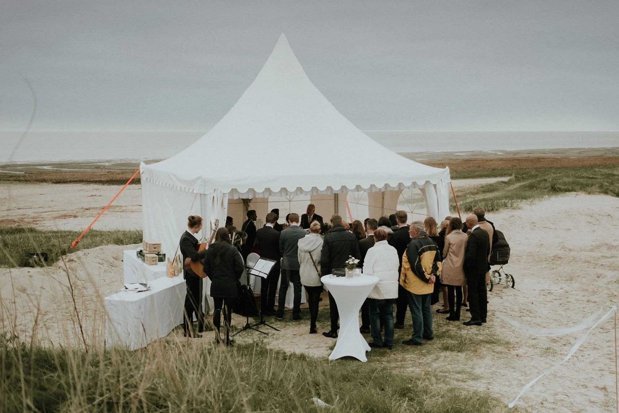 Gruppen von Menschen, die bei einer Veranstaltung unter einem weißen Zelt draußen am Strand versammelt sind.