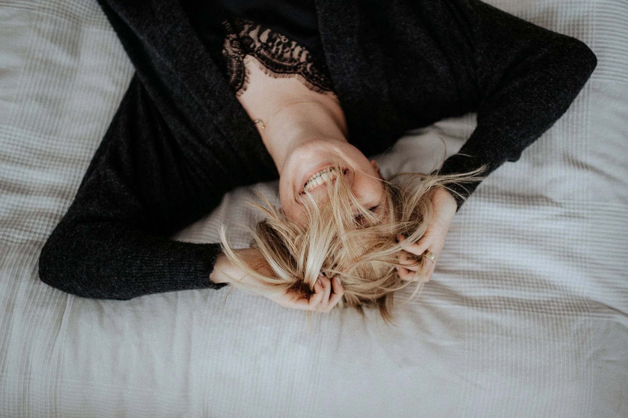 Eine lachende blonde Frau liegt auf einem Bett, umgeben von einem grauen und weißen Bettbezug, mit einem schwarzen Oberteil mit Spitze und einem hellen Unterhemd.