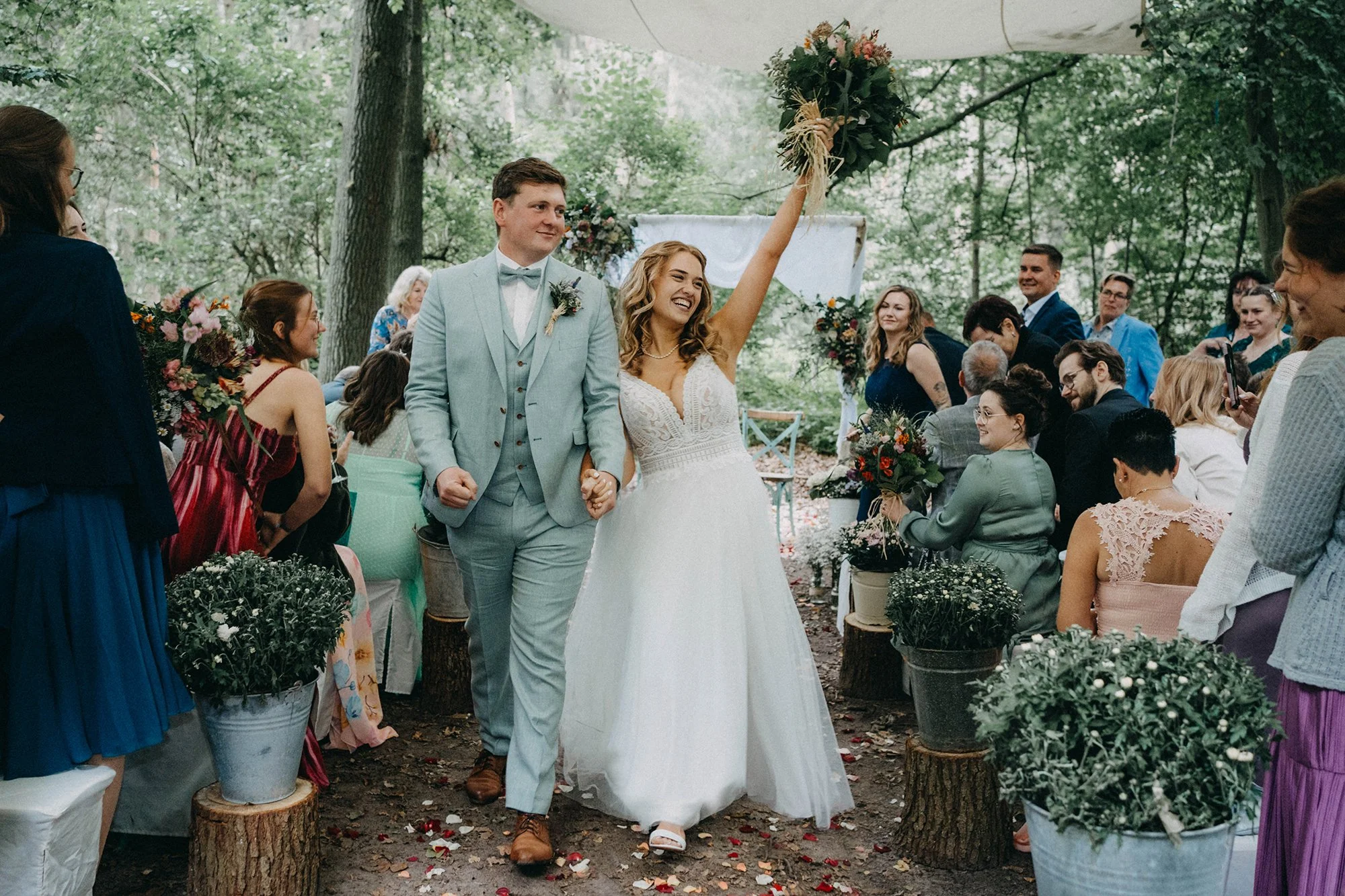 Ein glückliches Brautpaar bei einer Hochzeit im Freien, umgeben von Gästen, Blumen und Natur, während die Braut einen Blumenstrauß hochhält.