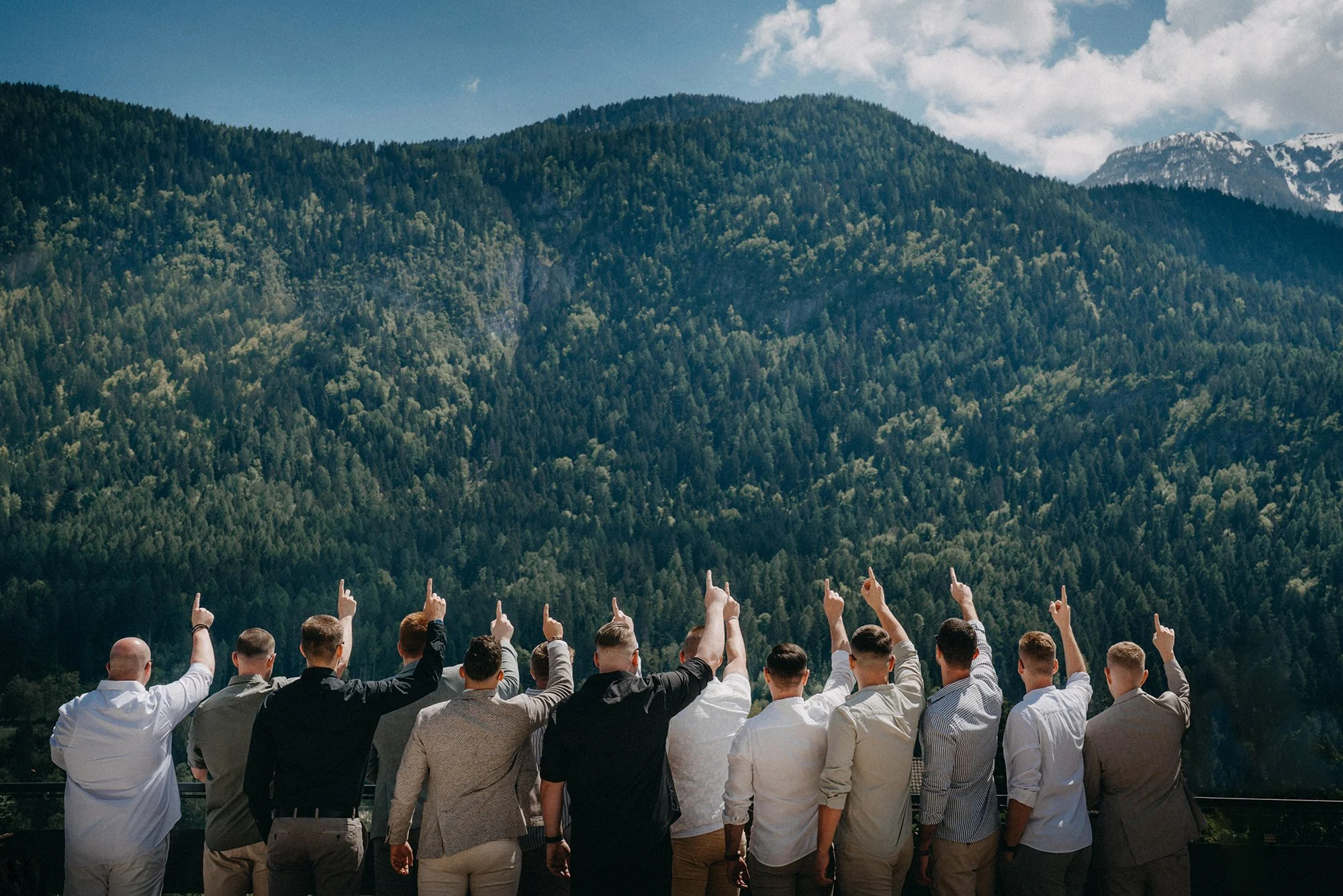 Eine Gruppe von Menschen steht im Vordergrund und zeigt nach oben, während im Hintergrund eine bewaldete Berglandschaft zu sehen ist. Es ist ein sonniger Tag mit einigen Wolken am Himmel.